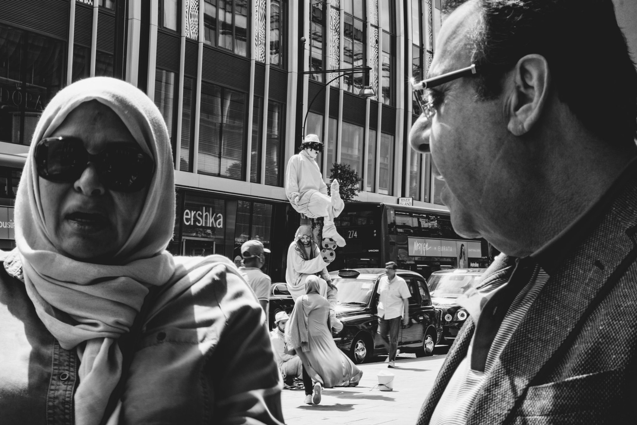 Czarno-białe zdjęcie uliczne z mimem na szczycie piramidy z ludzi, z przechodniami w tle i budynkami. Ujęcie z bliska na twarze dwóch osób.