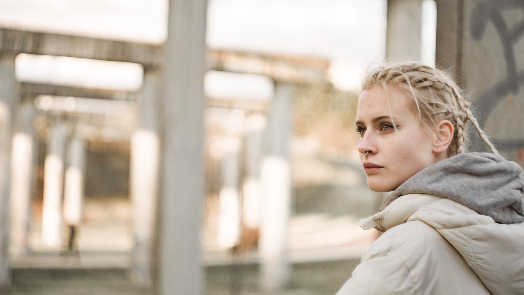 Portret młodej kobiety z blond warkoczami, w jasnej kurtce, z kolczykiem w nosie, na tle rozmytej, betonowej konstrukcji. Ujęcie z profilu, z delikatnym oświetleniem.
