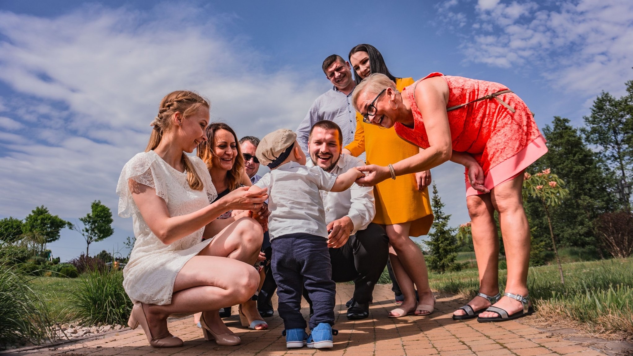 Uśmiechnięta rodzina otacza małego chłopca na brukowanej ścieżce w plenerze. Jasne ubrania, błękitne niebo i radosna atmosfera. Fotografia rodzinna w słoneczny dzień.