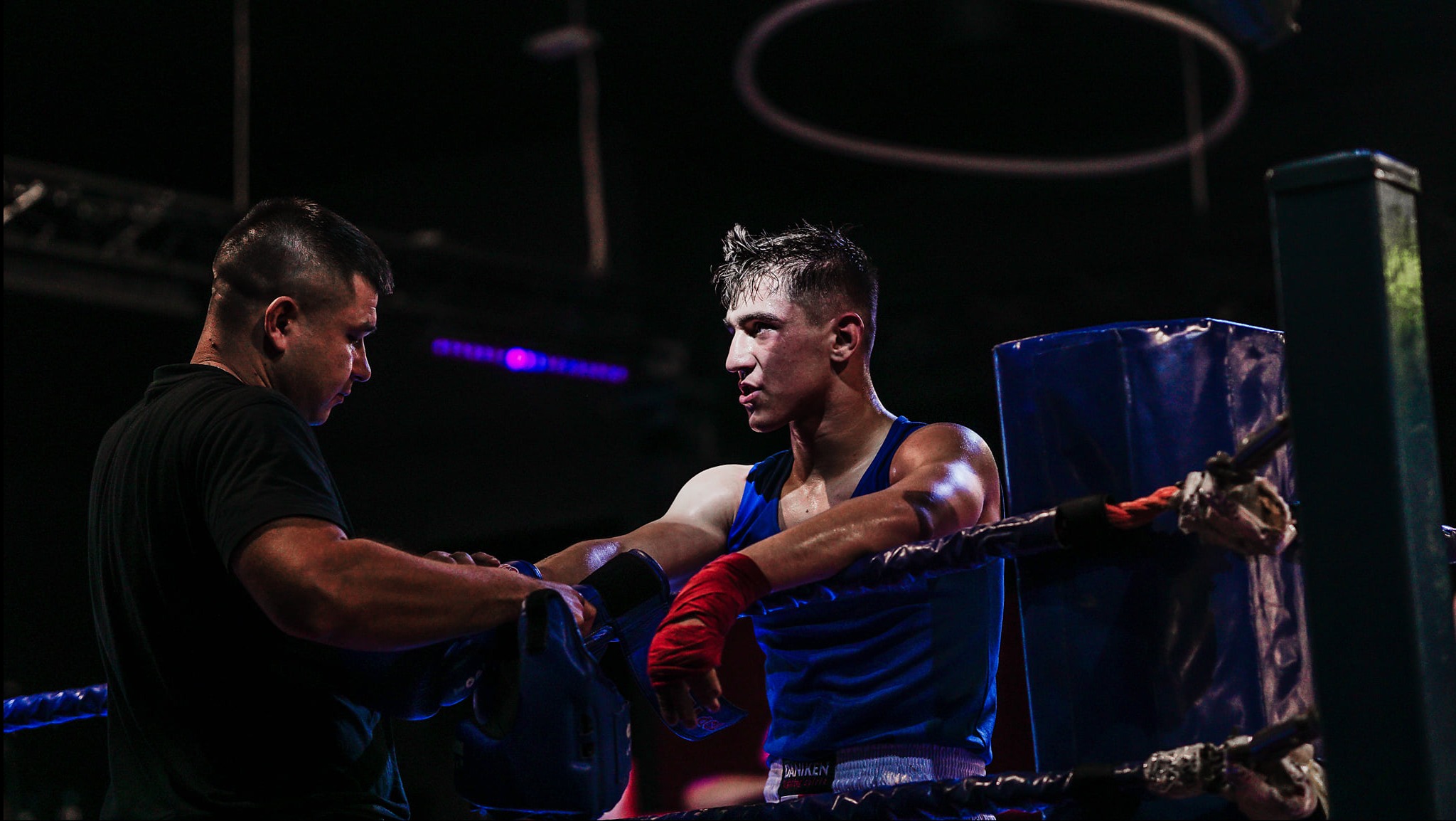 Bokser w niebieskim stroju opiera się o liny ringu, z trenerem obok, w tle ciemność i oświetlenie ringu. Dynamiczne ujęcie z zawodów bokserskich.