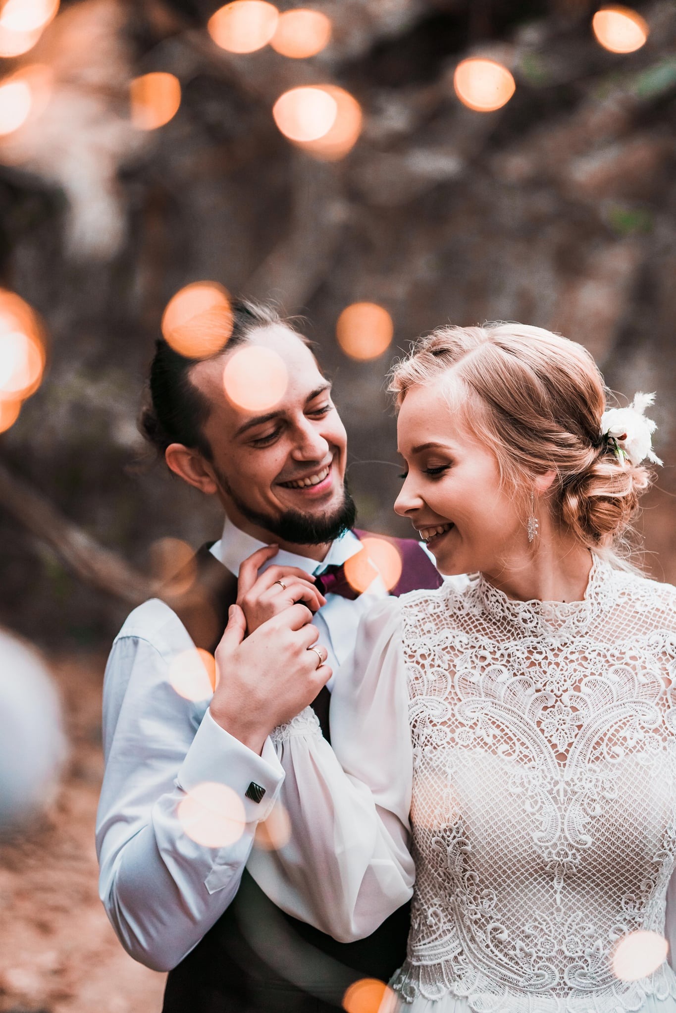 Uśmiechnięta para młoda w plenerze, panna młoda w koronkowej sukni, pan młody w muszce, ciepłe światło bokeh w tle, romantyczna atmosfera.