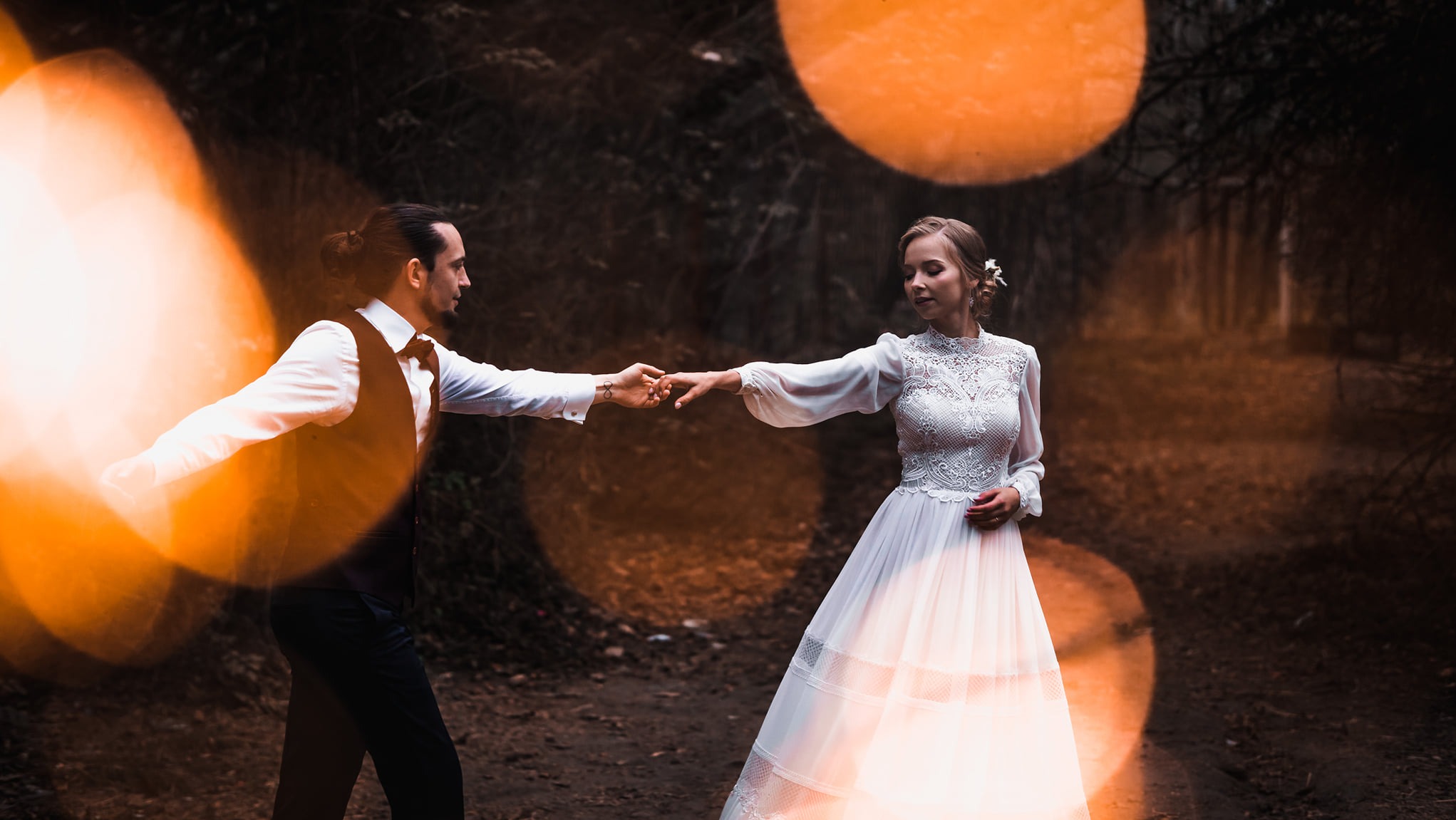 Para młoda w lesie, trzymająca się za ręce. Panna młoda w koronkowej sukni, pan młody w kamizelce i muszce. Bokeh w tle dodaje romantycznego nastroju.