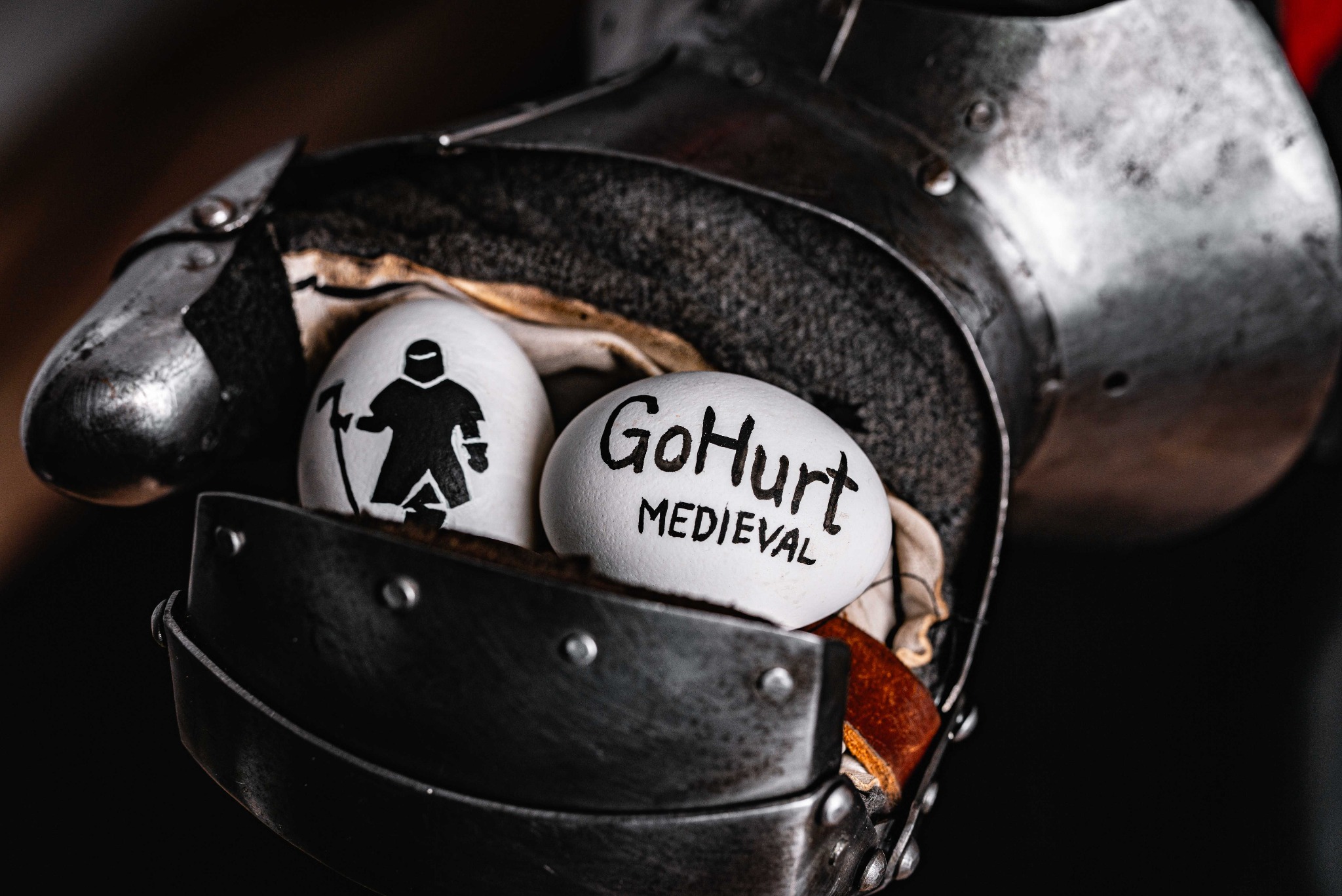 Dwa białe jajka z rysunkami i napisem 'GoHurt Medieval' w rękawicy zbroi rycerskiej. Nietypowe ujęcie, ciemne tło, ostre światło.
