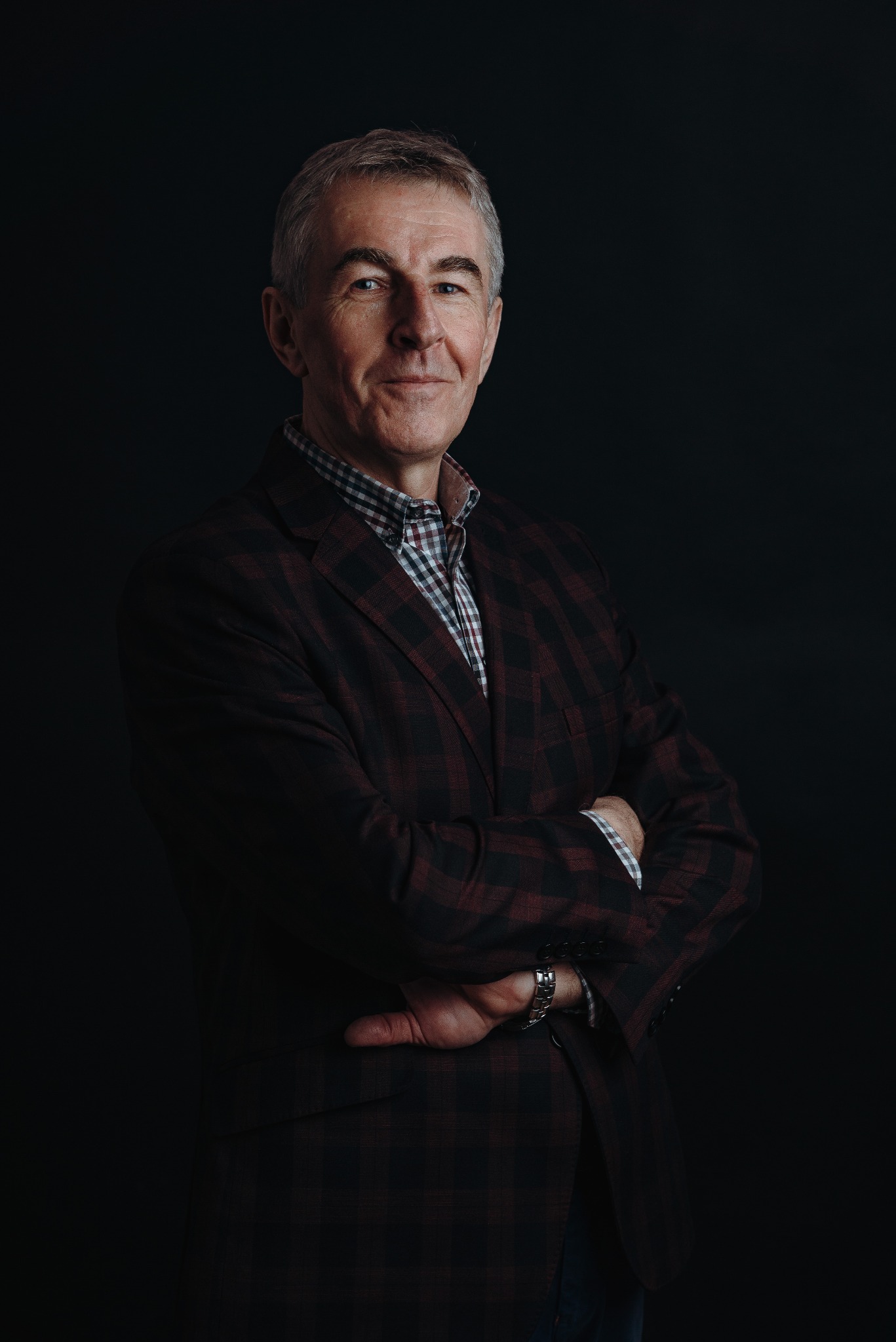 Portret biznesowy: Dojrzały mężczyzna w bordowej marynarce w kratę, skrzyżowane ręce, ciemne tło, profesjonalny wygląd, srebrny zegarek na nadgarstku.