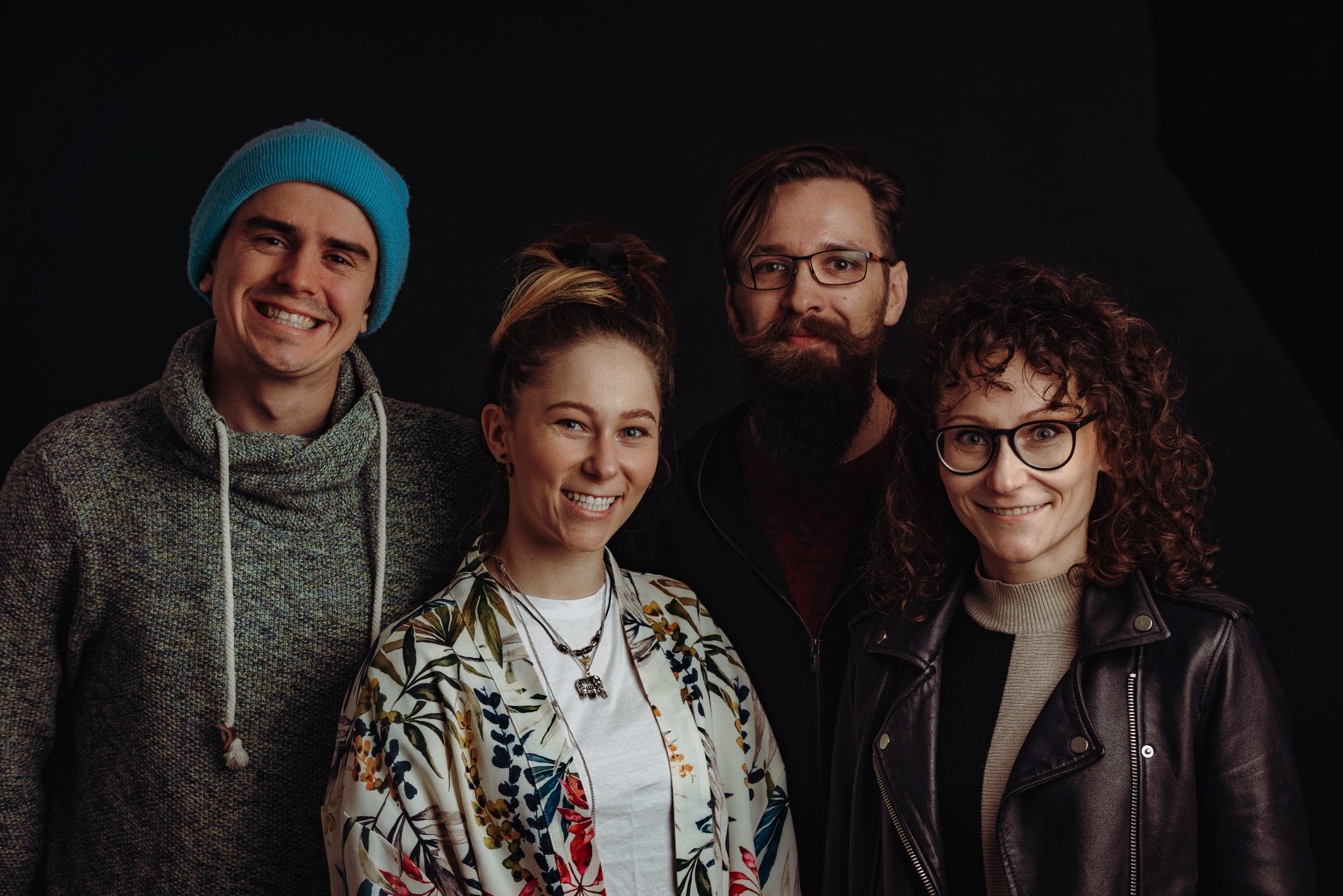 Portret grupowy czterech uśmiechniętych osób na ciemnym tle. Mężczyzna w niebieskiej czapce, kobieta w kwiecistej bluzce, mężczyzna z brodą i kobieta w skórzanej kurtce.