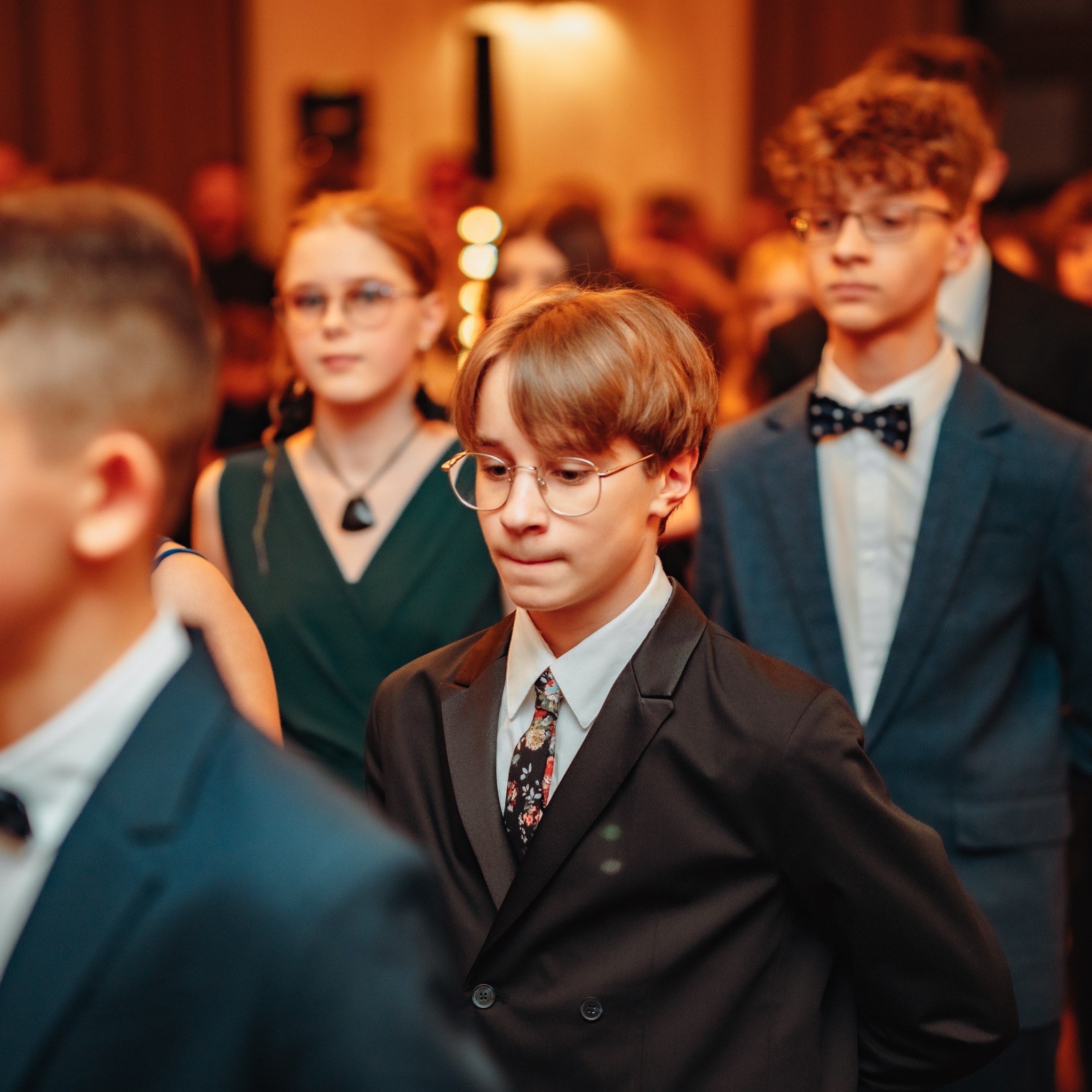 Młodzież w eleganckich strojach na tle rozświetlonego wnętrza. Chłopiec w czarnym garniturze i kwiecistym krawacie na pierwszym planie. Uczestnicy wydarzenia.