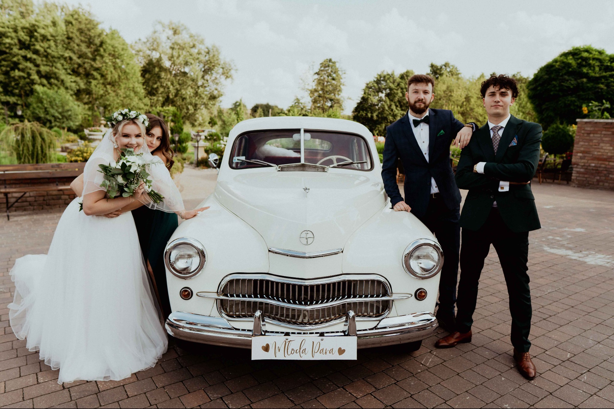 Uśmiechnięta panna młoda z bukietem i druhną pozuje obok białego, zabytkowego auta z napisem 'Młoda Para'. Obok stoją dwaj eleganccy mężczyźni w garniturach.