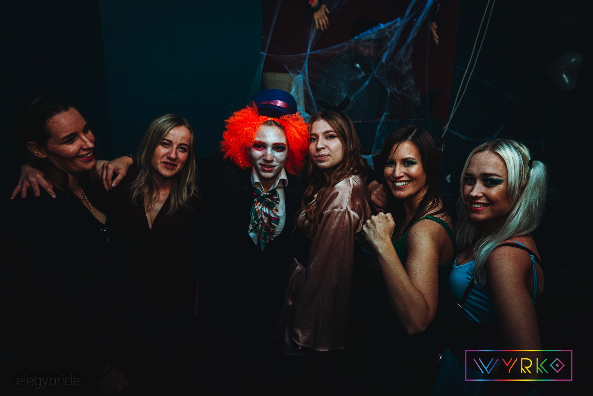 Grupa uśmiechniętych osób w kostiumach na imprezie tematycznej, w tle dekoracje halloweenowe. Jedna osoba w przebraniu Szalonego Kapelusznika.