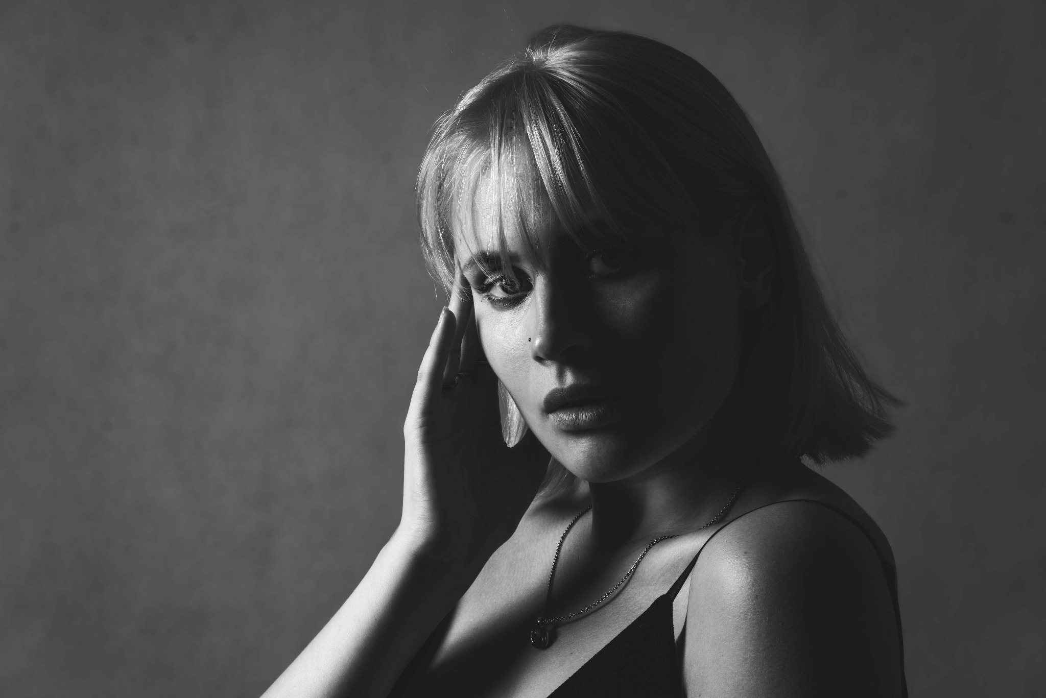 Czarno-biały portret kobiety z krótkimi blond włosami, zamyślonym spojrzeniem i delikatną biżuterią, oświetlony bocznym światłem.