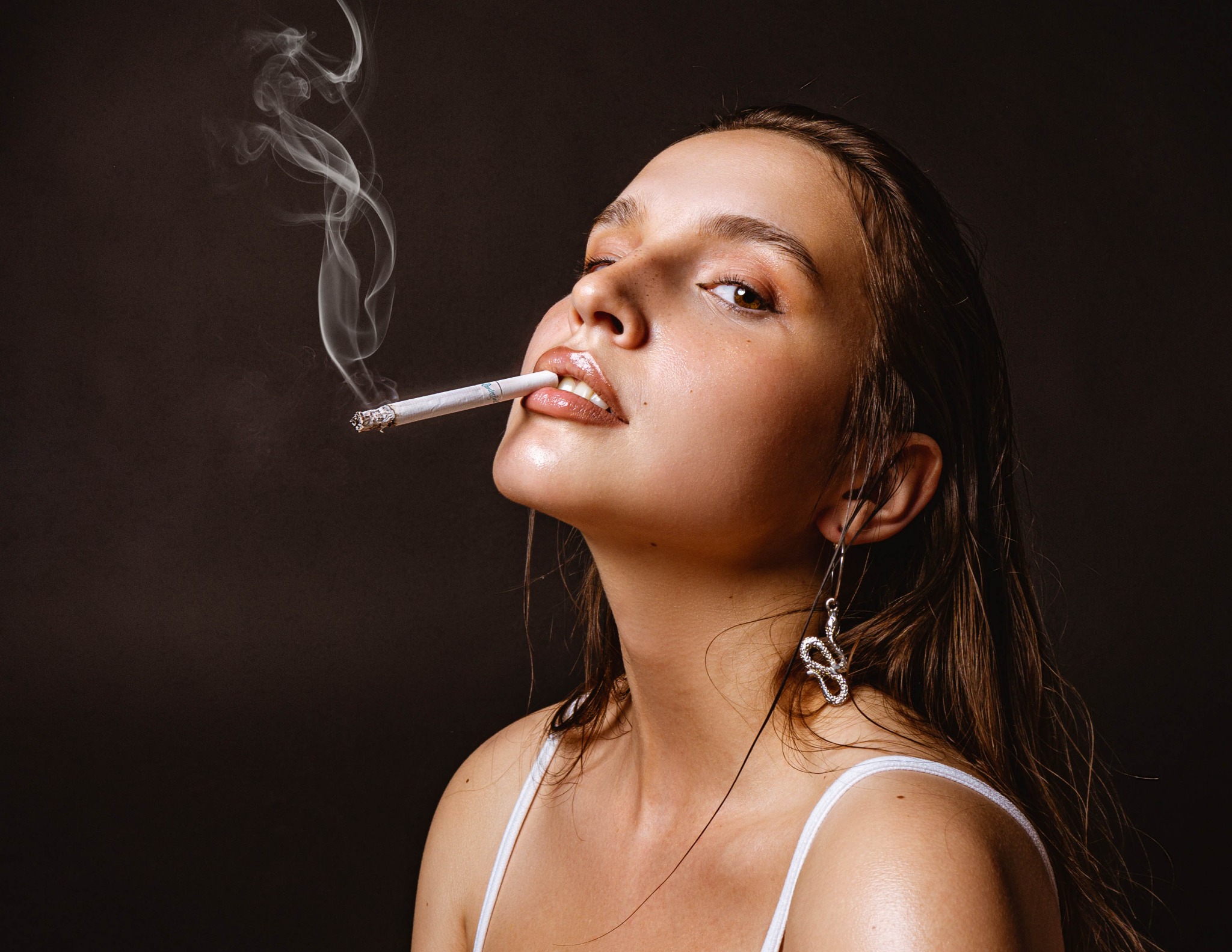 Portret kobiety z papierosem na ciemnym tle. Uwagę przyciągają błyszczące usta, mokre włosy i kolczyk w kształcie węża. Dym papierosowy tworzy subtelny efekt wizualny.