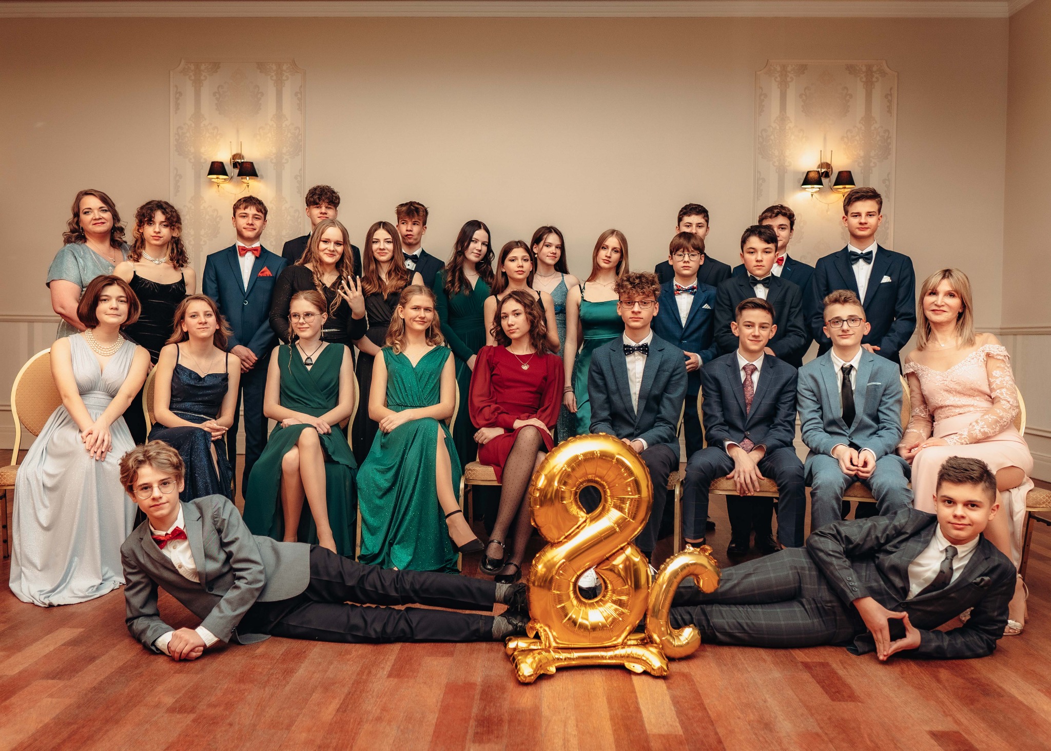 Grupowe, pozowane zdjęcie elegancko ubranych nastolatków i dorosłych na tle ściany z dekoracjami, z balonami w kształcie cyfr na podłodze.