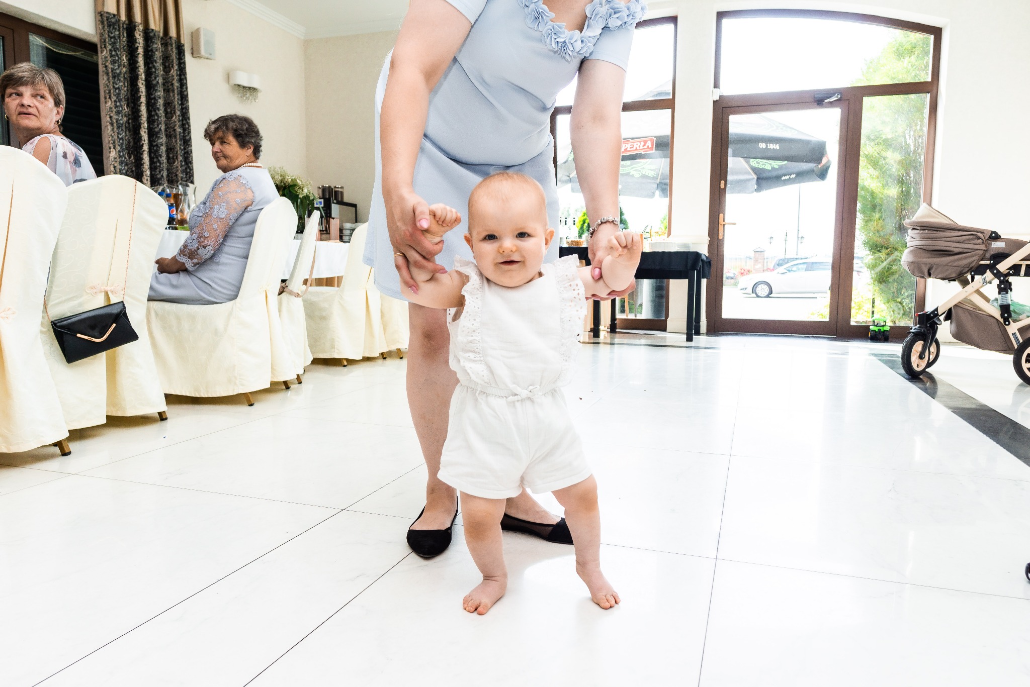 Uśmiechnięte dziecko w białym ubranku, uczące się chodzić, trzymane za ręce przez kobietę w jasnoniebieskiej sukience, w tle sala z gośćmi i wózkiem.