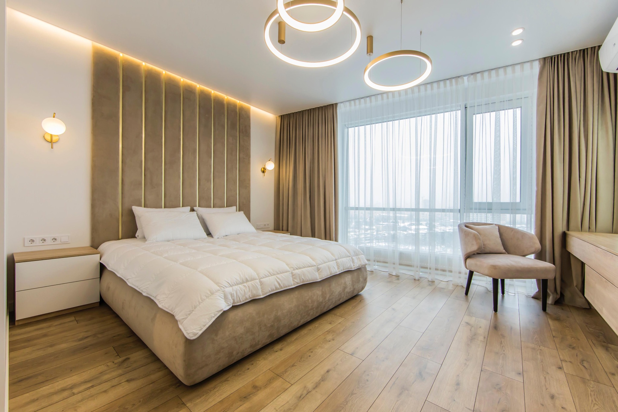 Nowoczesna sypialnia z podświetlanym wezgłowiem, okrągłymi lampami sufitowymi i drewnianą podłogą. Jasne wnętrze z dużym oknem i krzesłem.