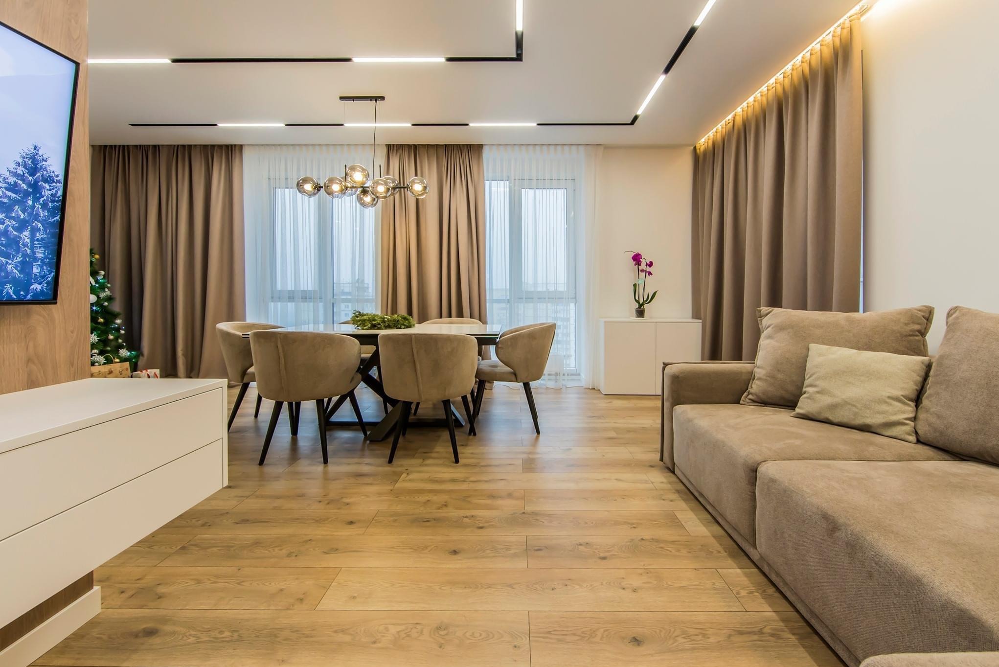 Jasne wnętrze salonu z drewnianą podłogą, beżową sofą i stołem jadalnym. Nowoczesne oświetlenie sufitowe i subtelne zasłony tworzą przytulną atmosferę.