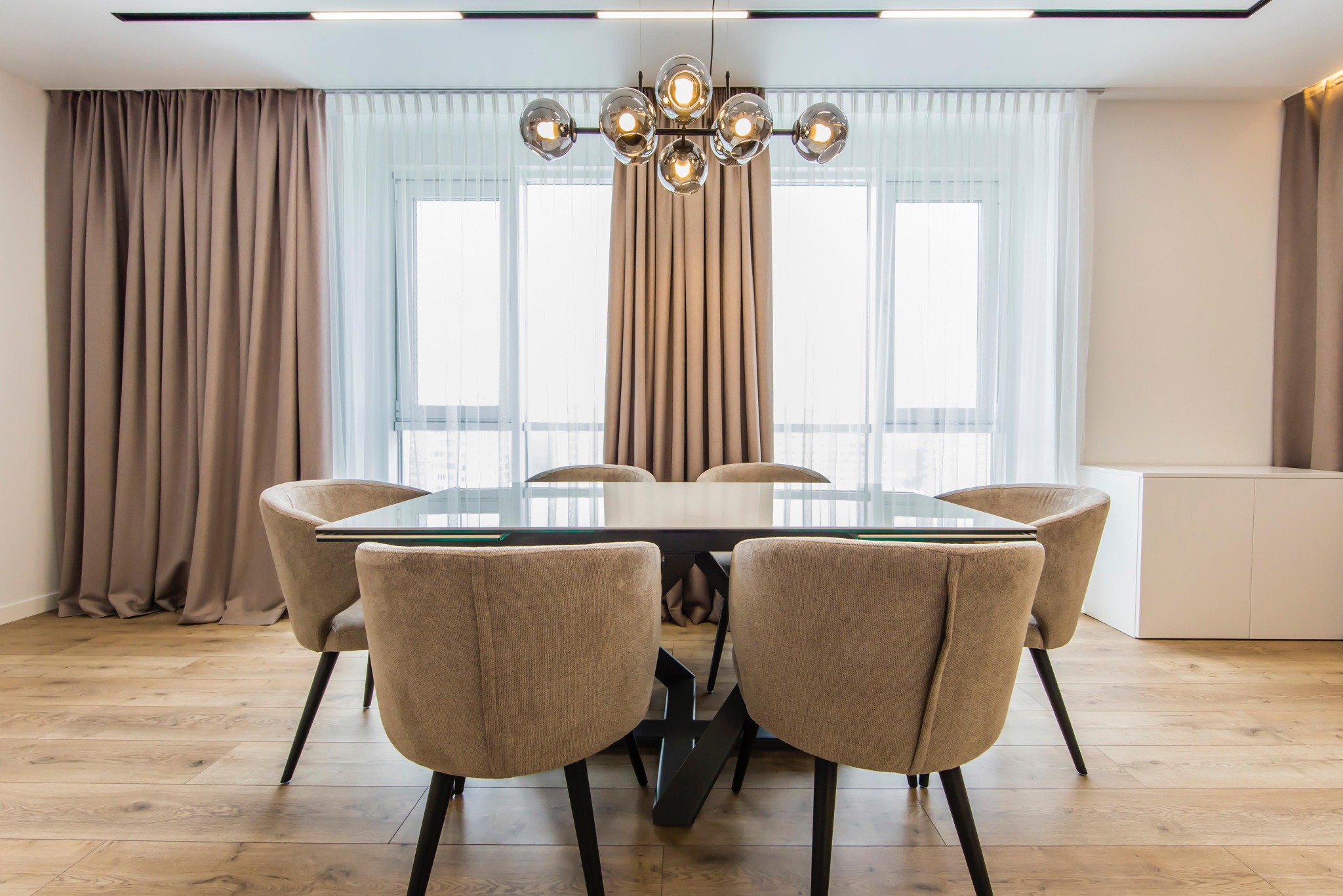 Jasne wnętrze jadalni z drewnianą podłogą, szklanym stołem i beżowymi krzesłami. W tle okno z zasłonami i nowoczesna lampa sufitowa.