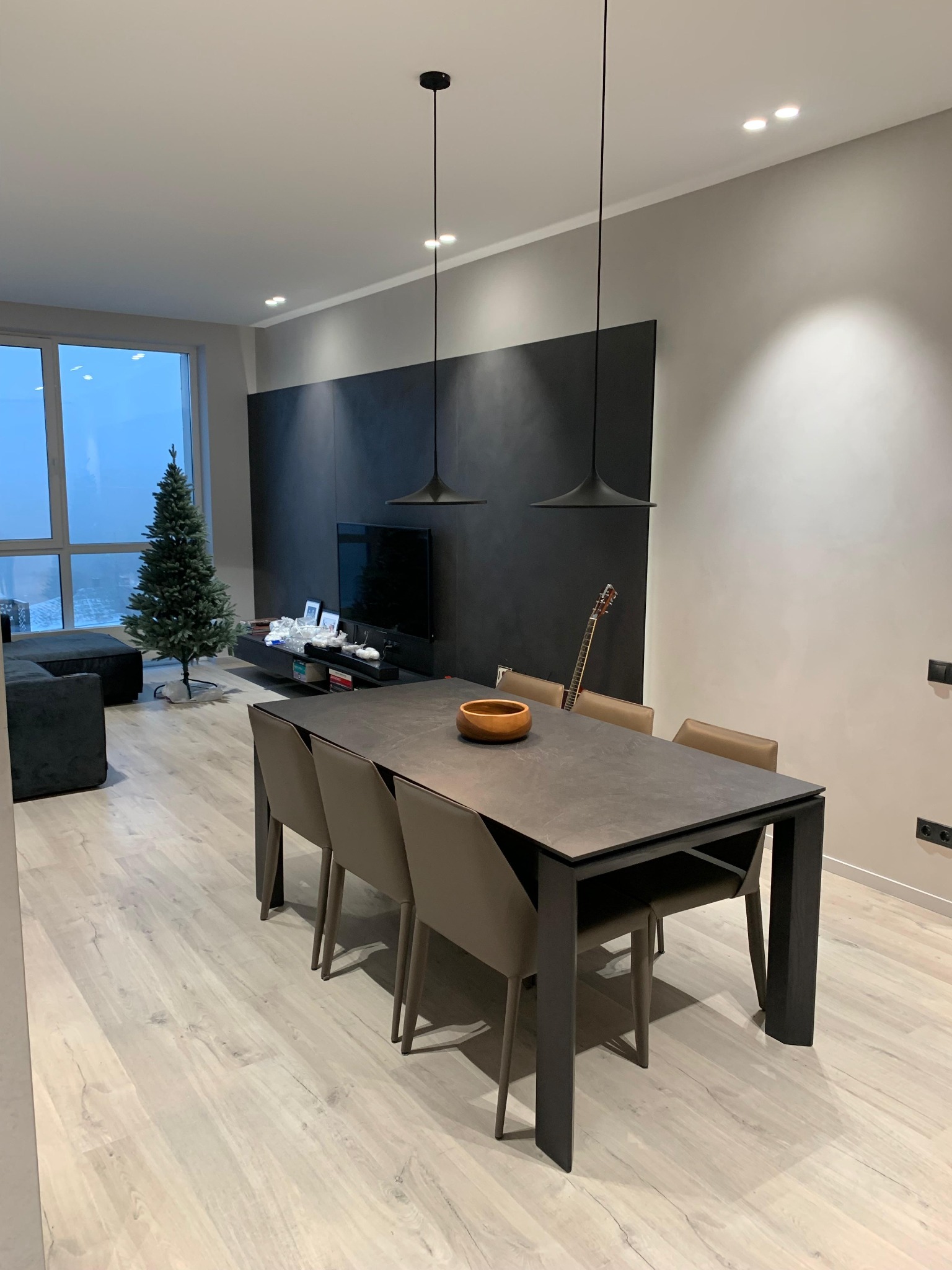 Nowoczesne wnętrze: stół z krzesłami, minimalistyczne lampy wiszące, ściana akcentowa w kolorze grafitu, parkiet w jasnym odcieniu drewna i choinka przy oknie.