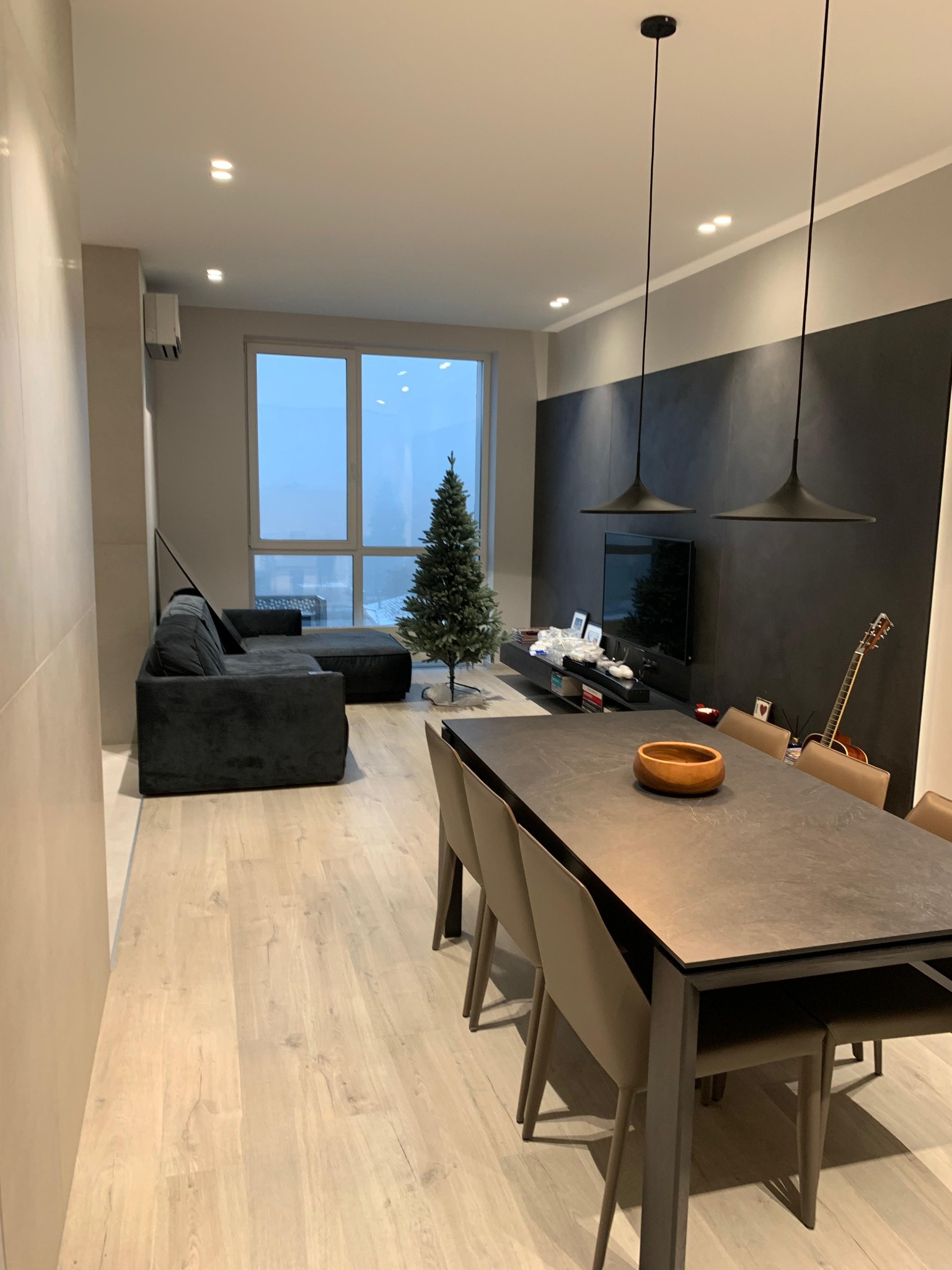 Nowoczesne wnętrze salonu z dużym oknem, szarą sofą narożną, stołem jadalnym z krzesłami i choinką. Minimalistyczne lampy wiszące nad stołem. Gitara oparta o ścianę.
