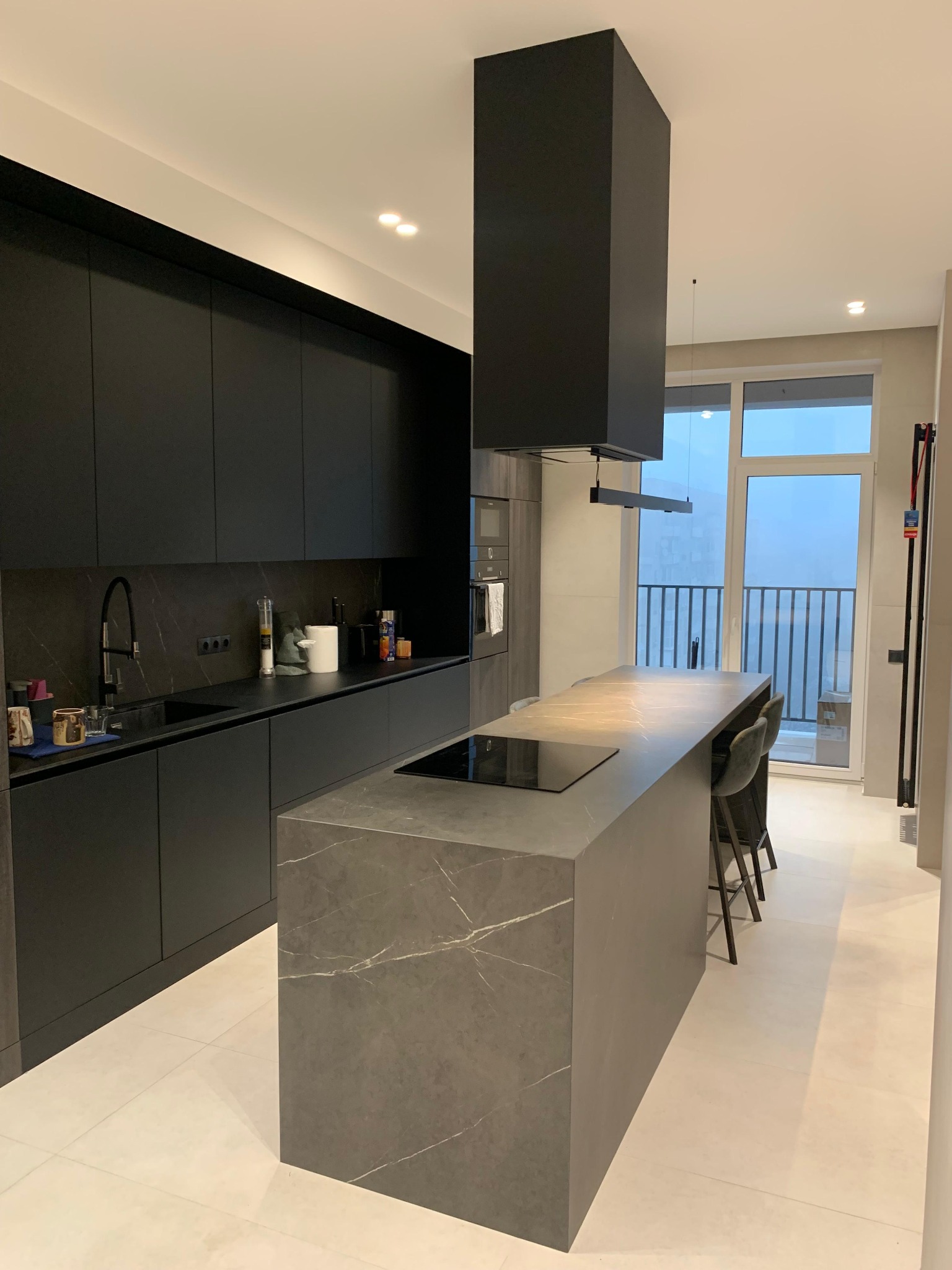 Nowoczesna kuchnia w ciemnej tonacji z wyspą i oknem balkonowym. Minimalistyczny design, matowe fronty szafek, marmurowy blat wyspy. Widok na balkon.