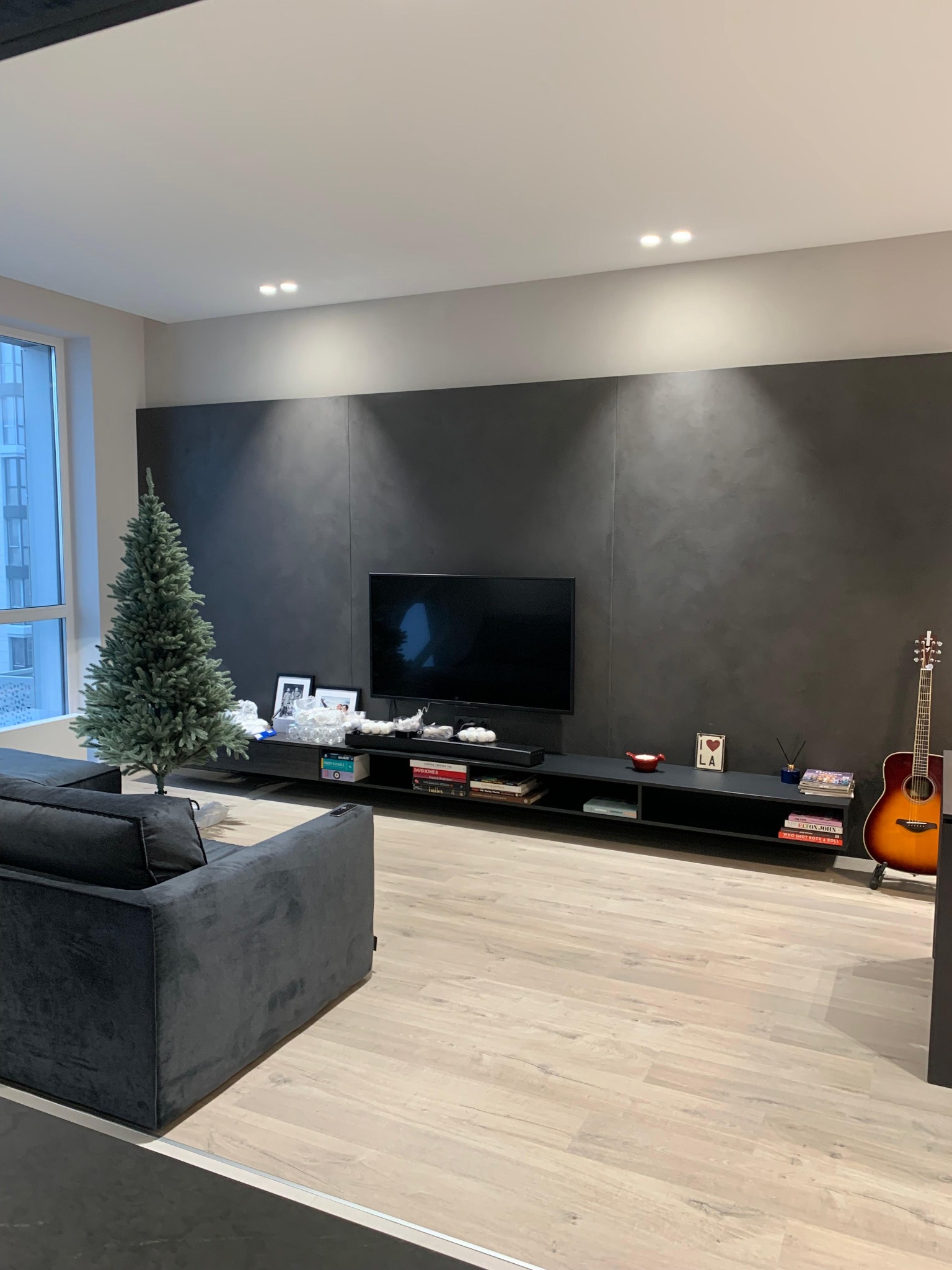 Nowoczesny salon z szarą ścianą, telewizorem, gitarą i choinką. Minimalistyczny design z drewnianą podłogą i czarnymi meblami. Duże okno z widokiem na miasto.