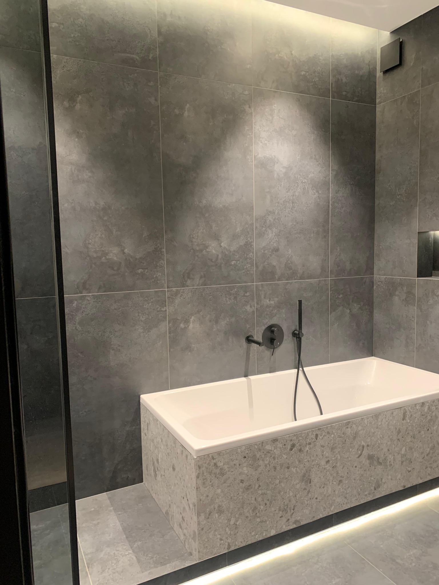 Nowoczesna łazienka z wanną obłożoną szarą mozaiką, grafitowe płytki na ścianach, czarna armatura i subtelne oświetlenie LED pod wanną.