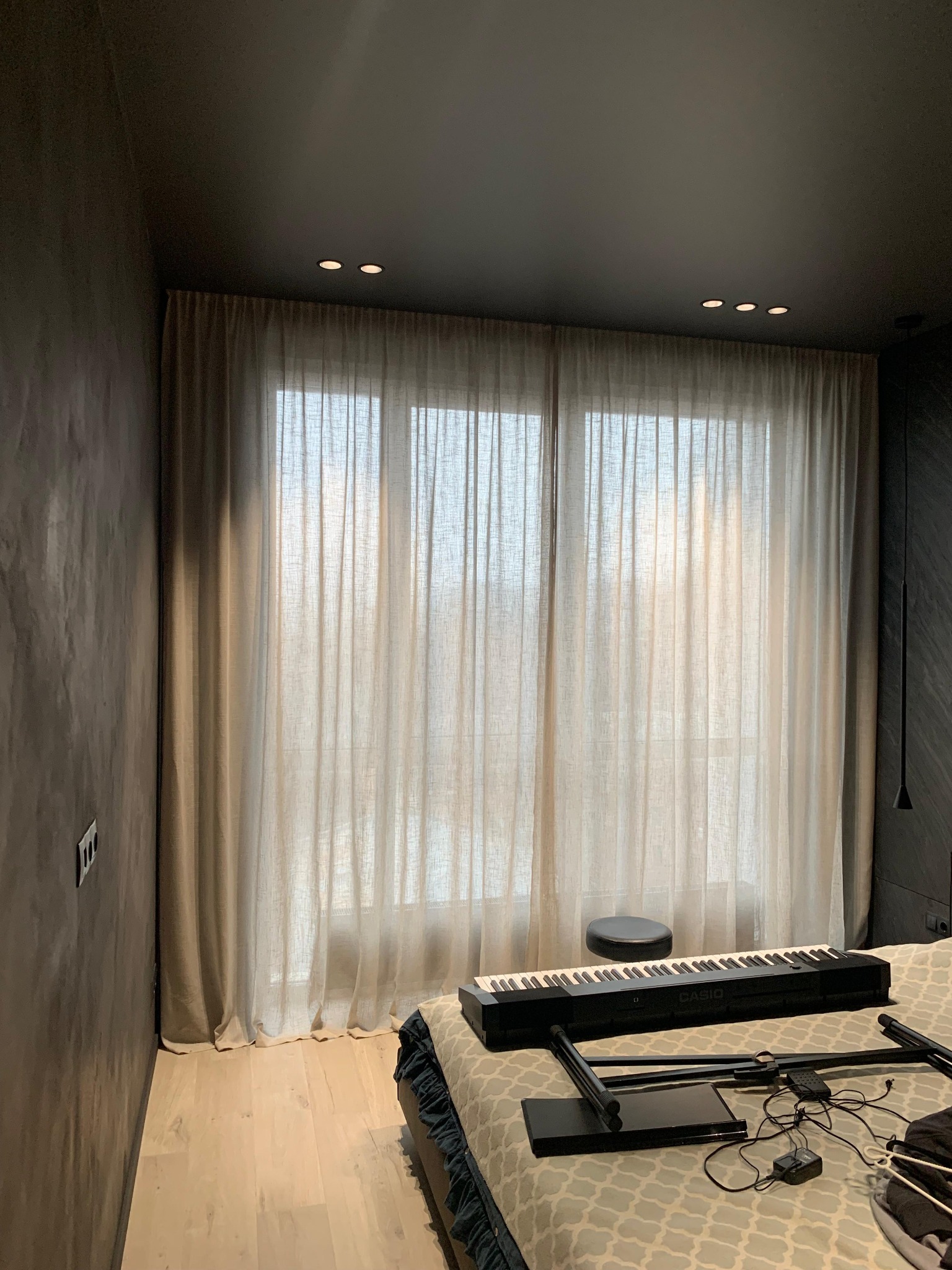 Beżowe, długie zasłony w nowoczesnym wnętrzu z ciemnymi ścianami i drewnianą podłogą. Widoczne oświetlenie sufitowe i fragment pianina na łóżku.