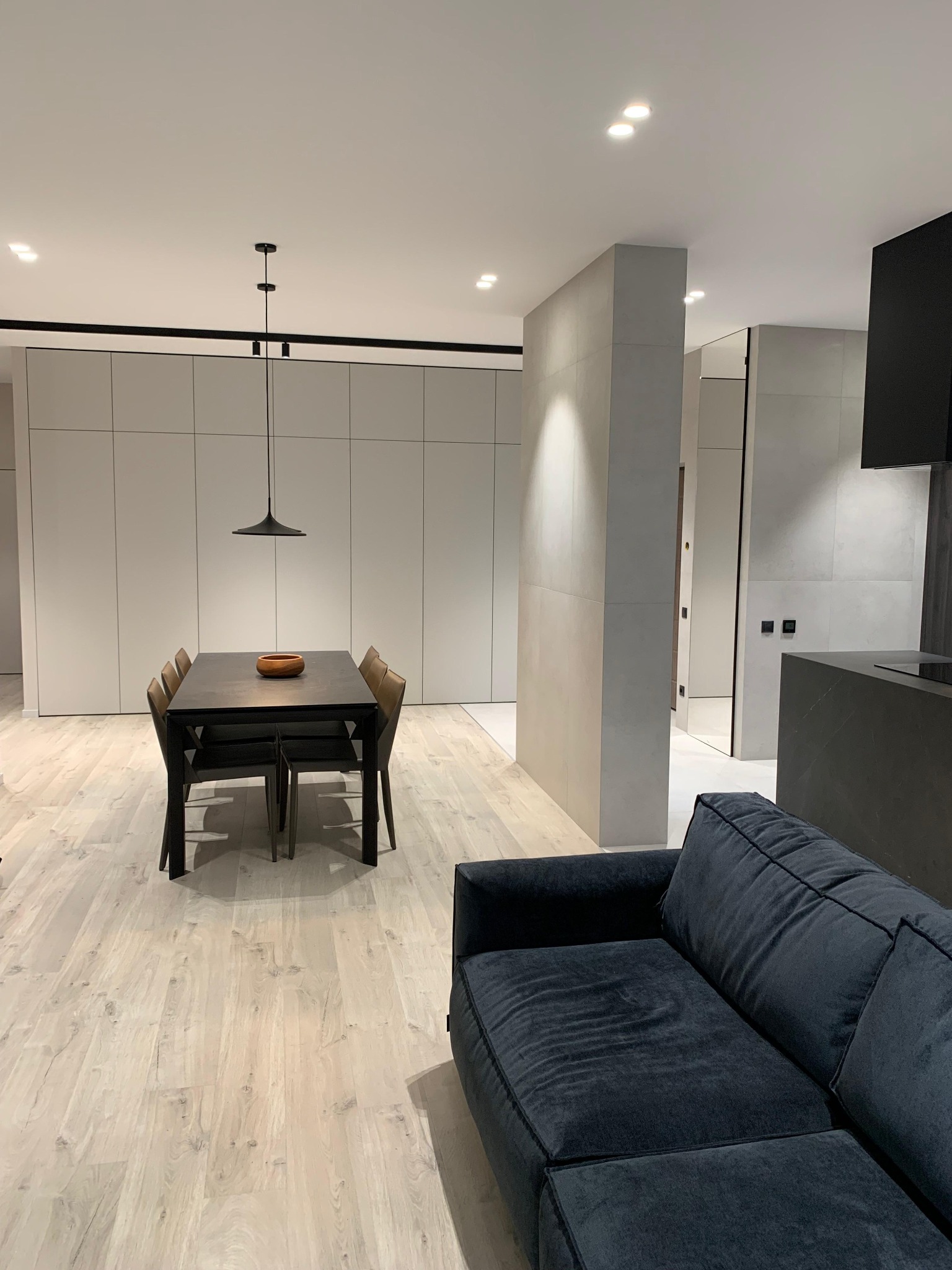 Nowoczesne wnętrze: stół z krzesłami, minimalistyczna lampa, zabudowa ścienna i fragment sofy. Jasna podłoga i stonowana kolorystyka tworzą spójną całość.