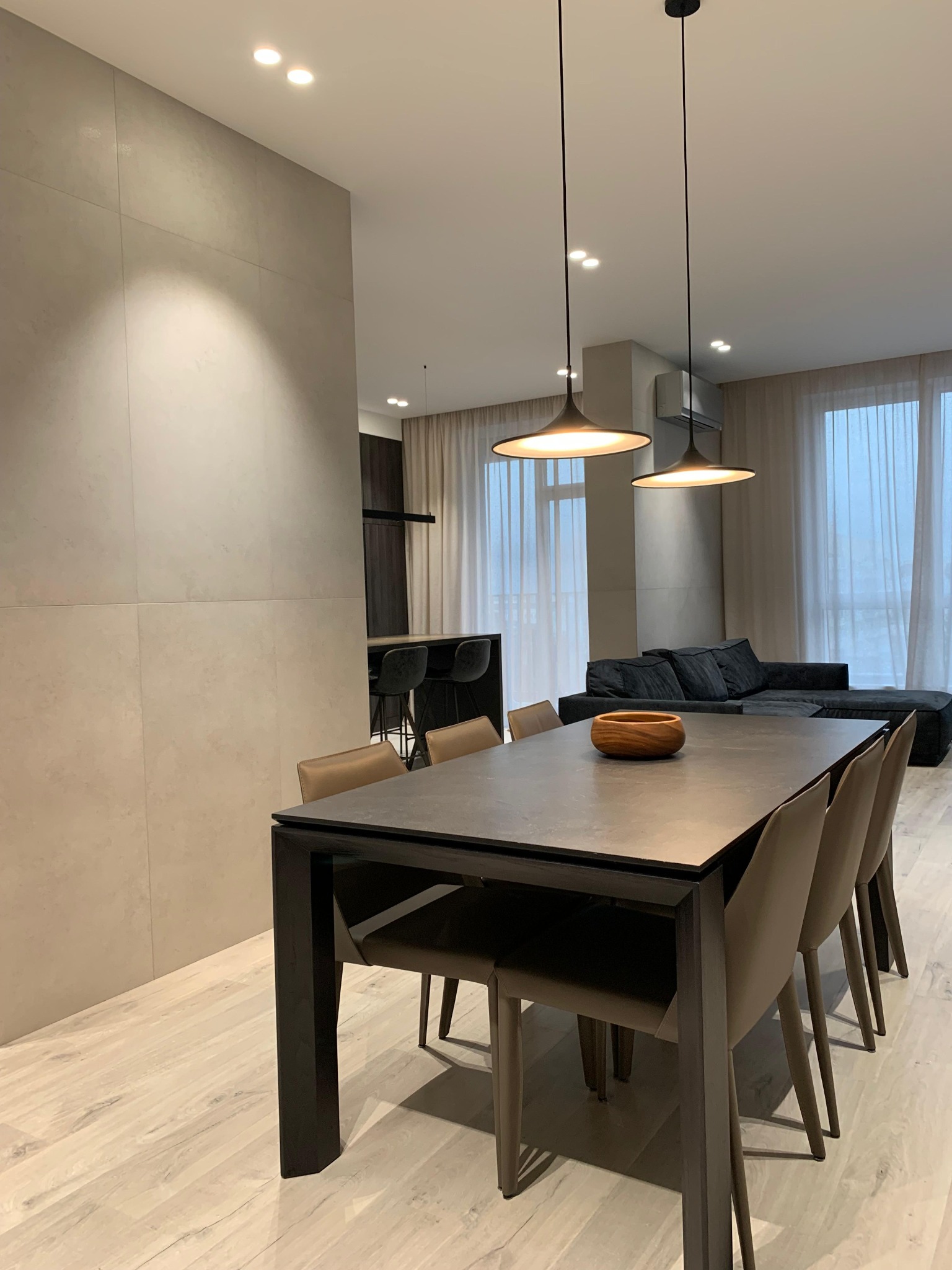 Nowoczesne wnętrze: stół z krzesłami w odcieniach szarości i beżu, minimalistyczne lampy wiszące, ściana wykończona płytami imitującymi beton, jasna podłoga.