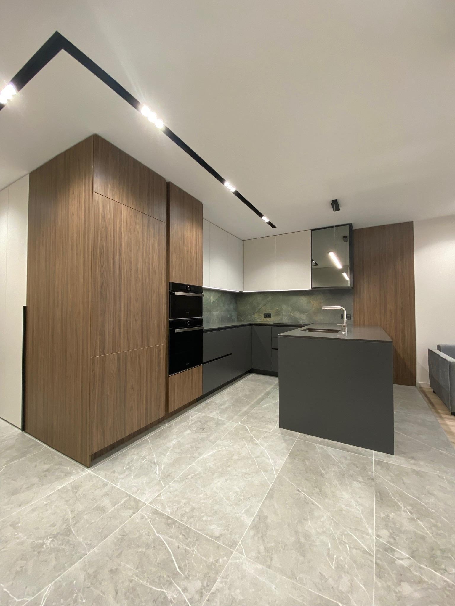Nowoczesna kuchnia z wyspą, drewnianymi frontami szafek i marmurową podłogą. Minimalistyczne oświetlenie sufitowe podkreśla elegancję wnętrza.