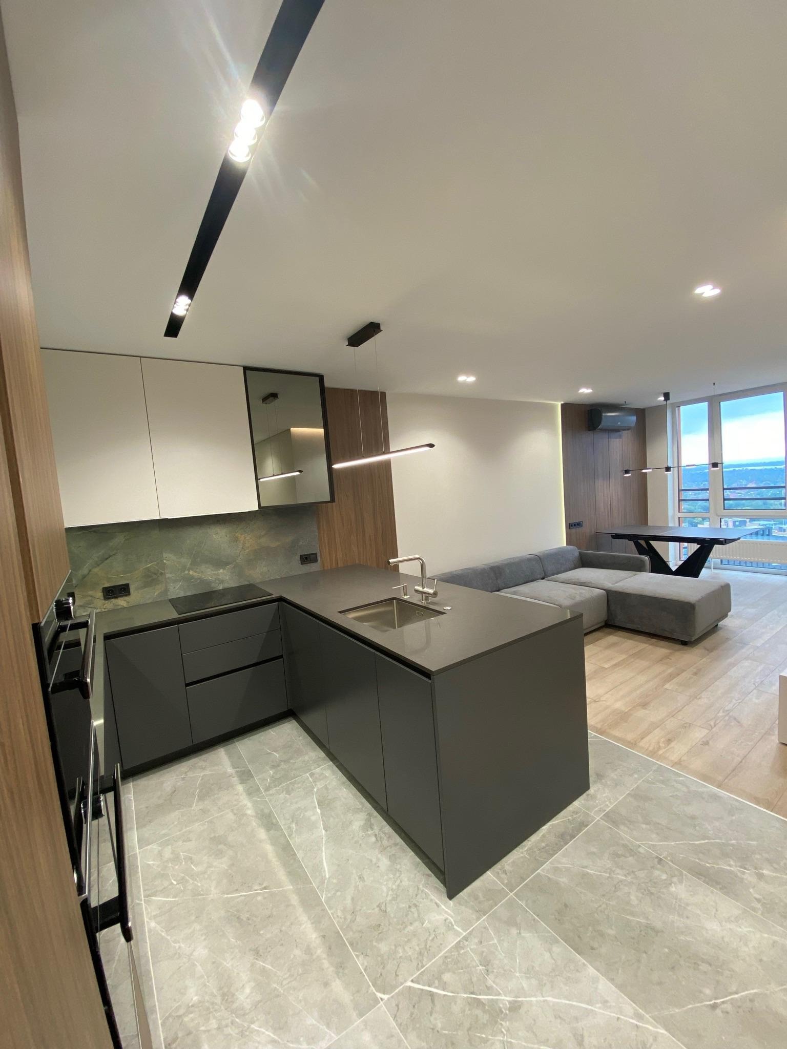 Nowoczesne wnętrze: szara kuchnia z wyspą, marmurowe płytki, drewniane akcenty, szara sofa i duże okna z widokiem na miasto. Minimalistyczny design z dbałością o detale.