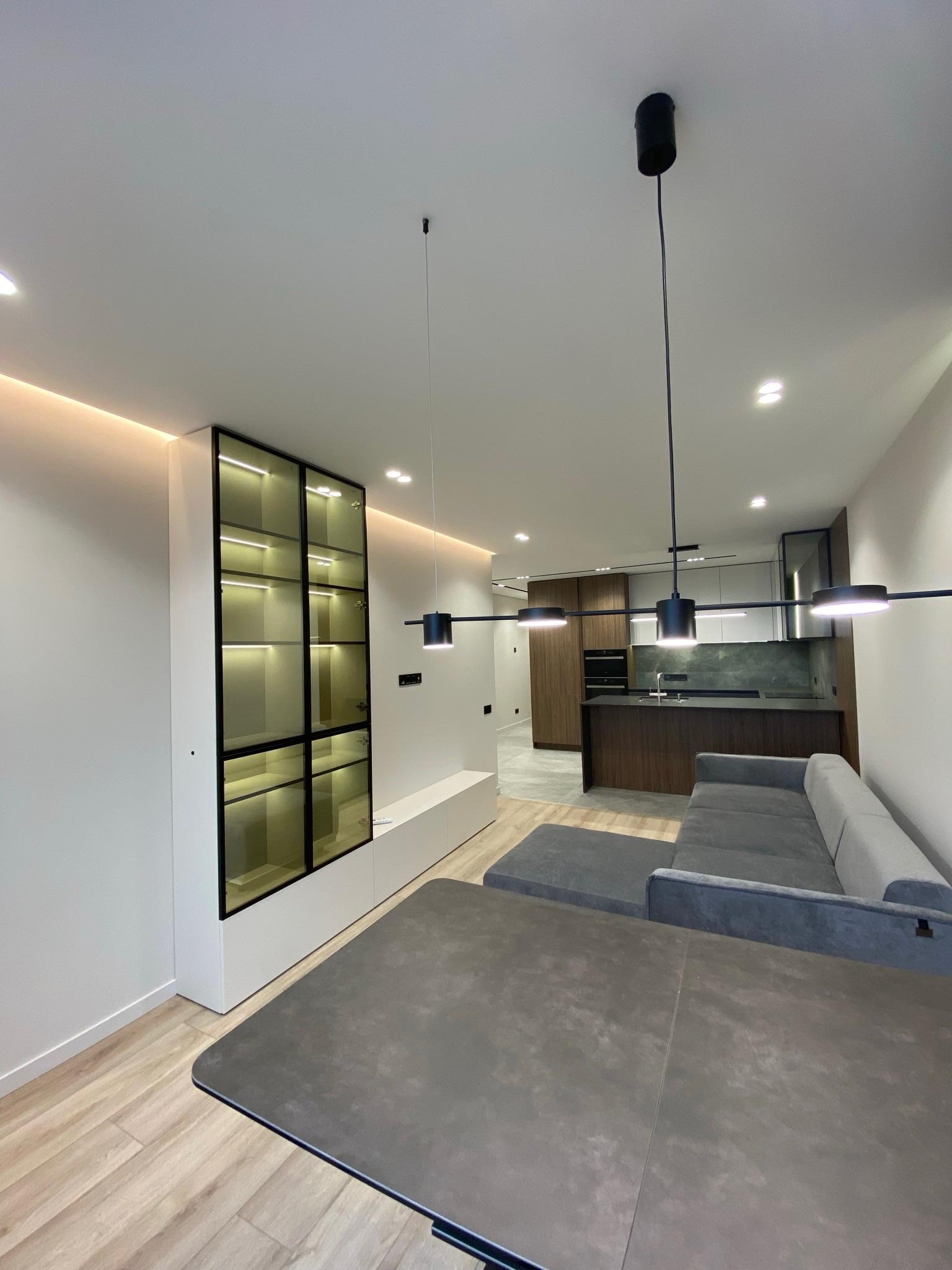 Nowoczesne wnętrze: szary narożnik, stół z blatem imitującym beton, przeszklona witryna z podświetleniem i otwarta kuchnia w tle. Minimalistyczne lampy.