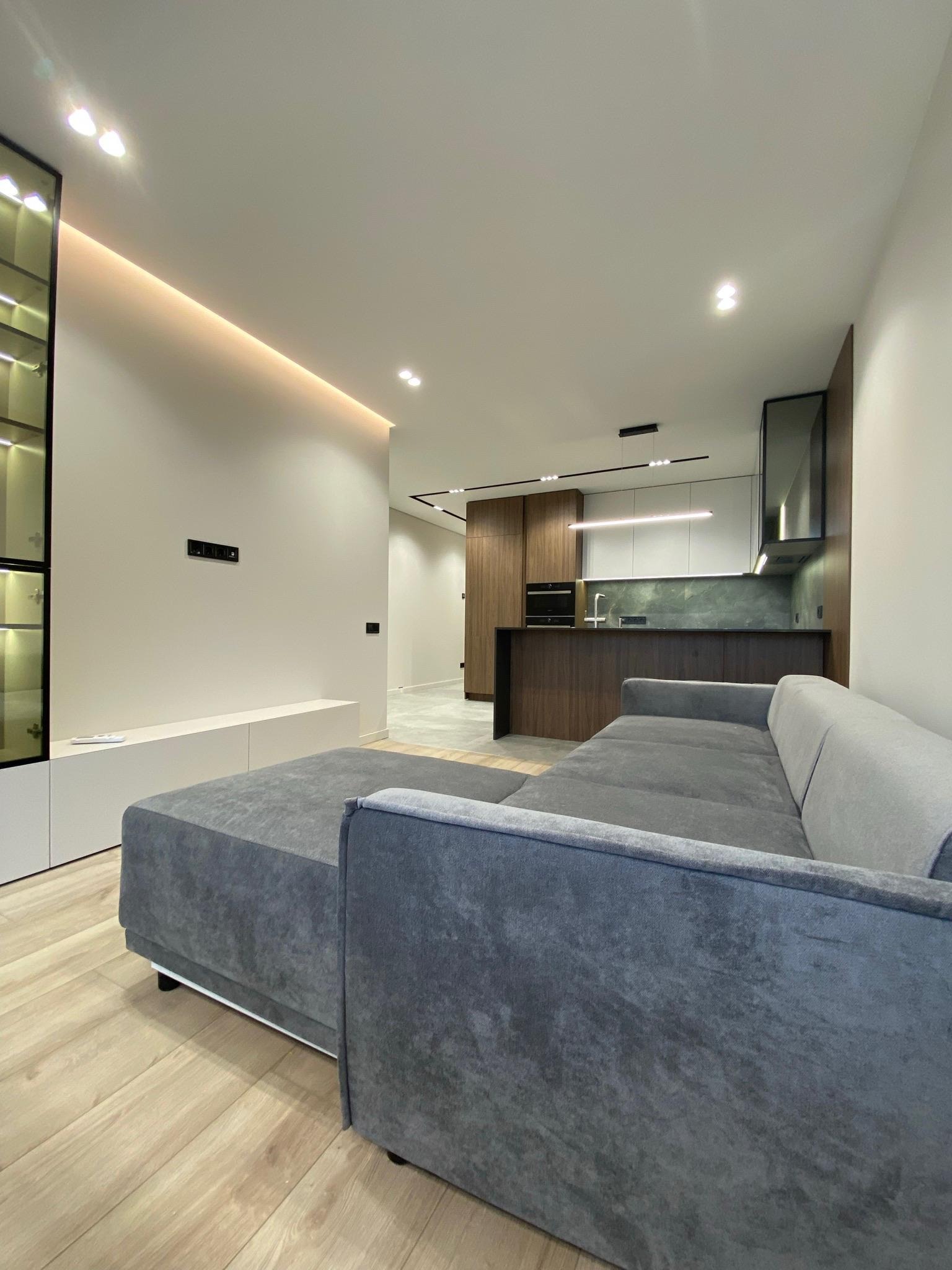 Nowoczesne wnętrze: szara sofa narożna, drewniana podłoga, oświetlenie LED i zabudowa kuchenna z elementami drewna. Minimalistyczny design.