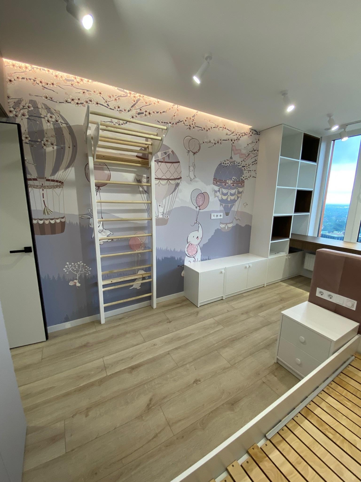 Pokój dziecięcy z drewnianą drabinką gimnastyczną na tle ściany z motywem balonów i zwierząt. Jasne drewniane panele podłogowe i białe meble.