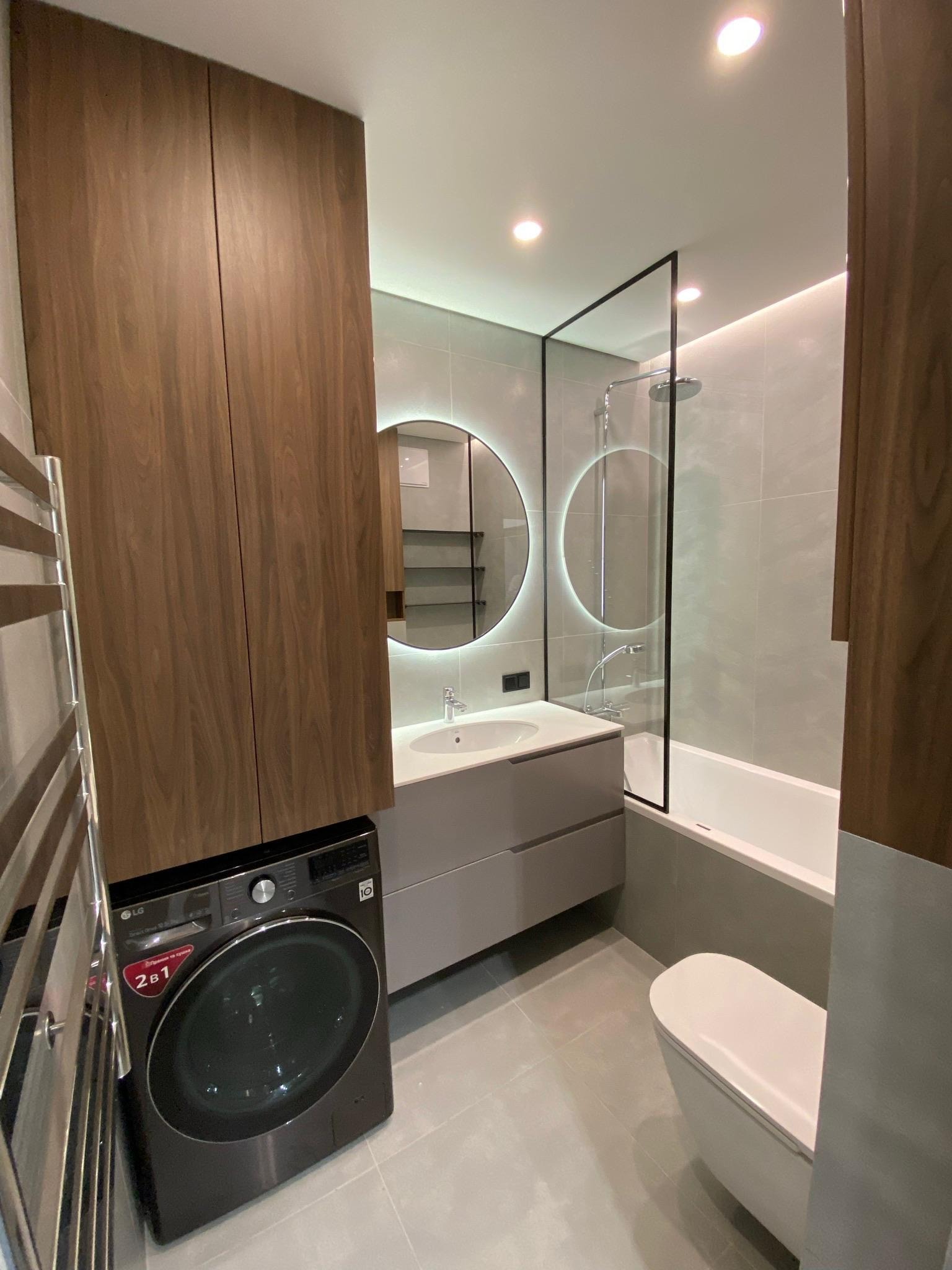 Nowoczesna łazienka z pralką pod zabudową, okrągłe lustro z podświetleniem LED, szklana kabina prysznicowa i minimalistyczna toaleta w odcieniach szarości i brązu.