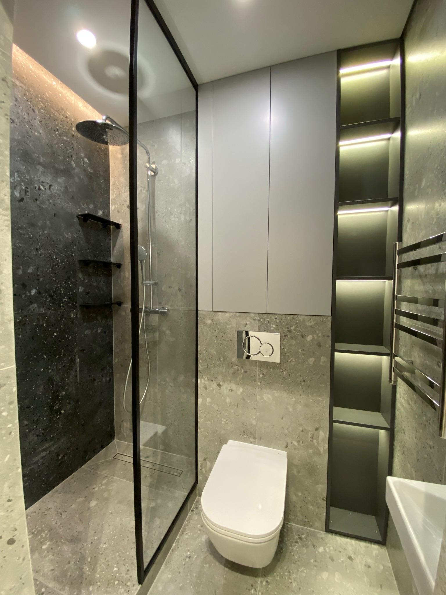 Nowoczesna łazienka z kabiną prysznicową typu walk-in, toaletą i podświetlanymi półkami wnękowymi. Szare płytki imitujące kamień na ścianach i podłodze.
