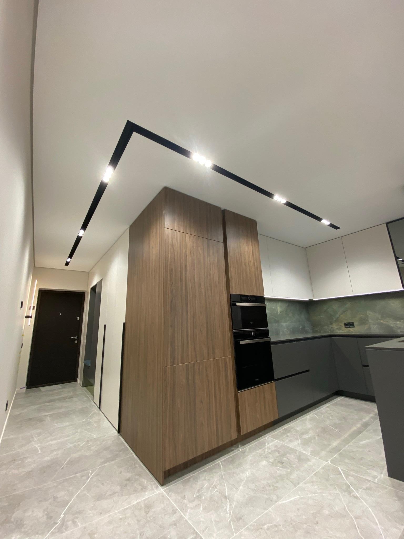 Nowoczesna kuchnia z drewnianymi i szarymi szafkami, marmurową podłogą i czarnym oświetleniem sufitowym. Widok na drzwi wejściowe.