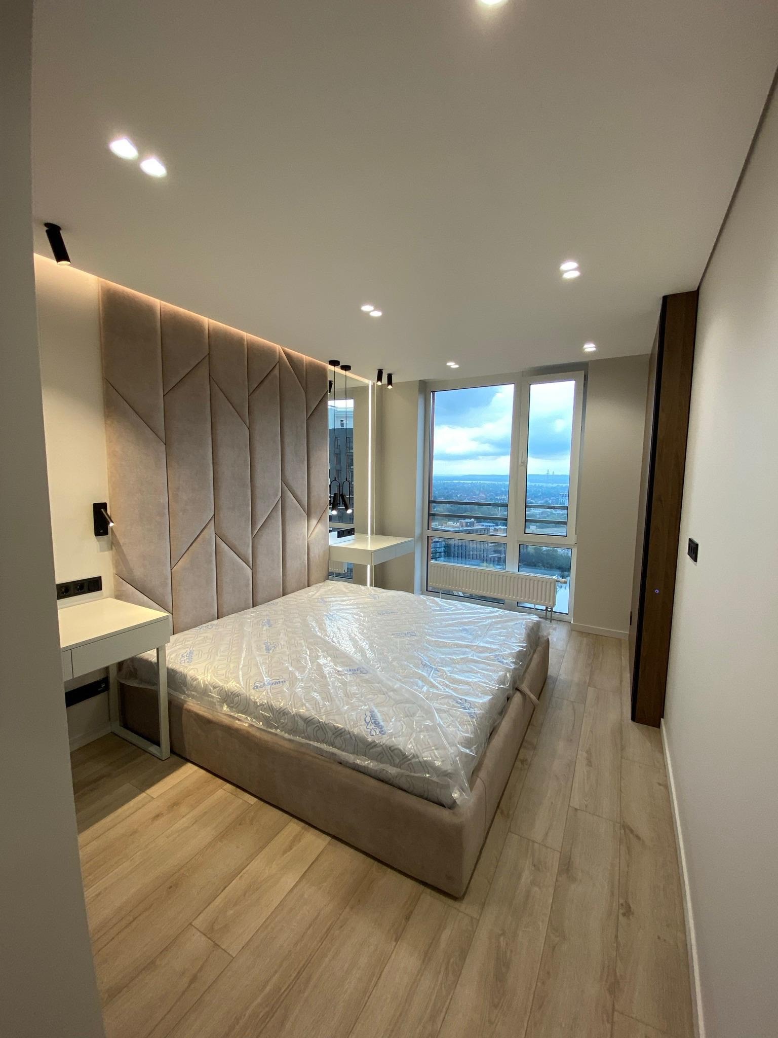 Nowoczesna sypialnia z łóżkiem z tapicerowanym zagłówkiem w geometryczne wzory, jasnymi panelami podłogowymi i dużym oknem z widokiem na miasto.