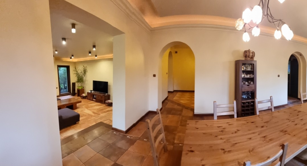 Jasne wnętrze domu z łukami, widok na salon z telewizorem i rośliną doniczkową. Na pierwszym planie drewniany stół i krzesła. Ściany w odcieniach beżu.