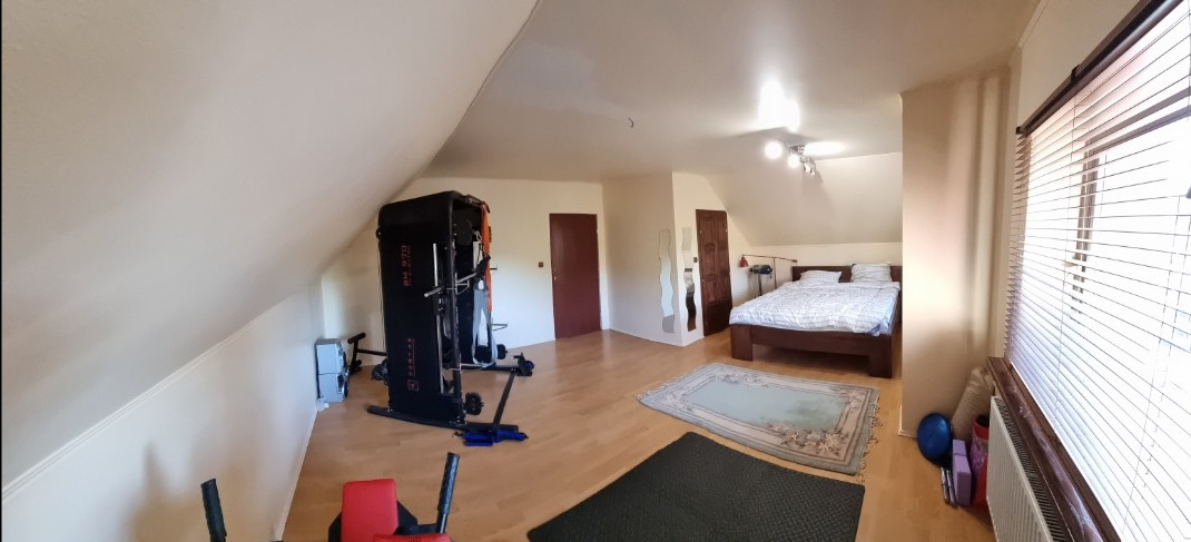 Jasne poddasze ze skośnym sufitem, urządzone jako sypialnia i domowa siłownia. Widoczne łóżko, dywany, maszyna do ćwiczeń i okno z żaluzjami.