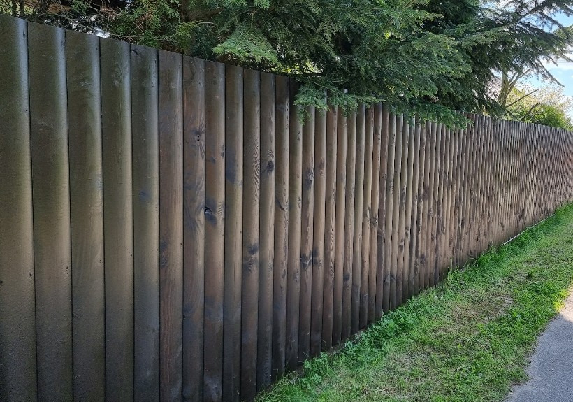Pomazany, ciemnobrązowy płot z okrąglaków, świeżo po malowaniu, z widocznymi smugami farby, otoczony zielenią i drzewami w tle.