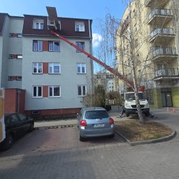 Fast-Up usługi podnośnikiem - Czerwony podnośnik koszowy ustawiony przy bloku mieszkalnym, z wysięgnikiem sięgającym dachu. Zaparkowane samochody i drzewo w tle. Dzień słoneczny.