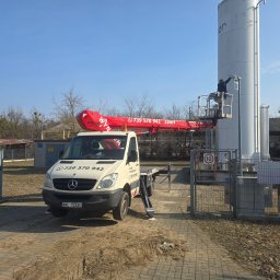 Fast-Up usługi podnośnikiem - Pracownik na podnośniku koszowym, w trakcie inspekcji lub naprawy wysokiego, białego zbiornika przemysłowego. Czerwony wysięgnik z numerem telefonu. Błękitne niebo w tle.