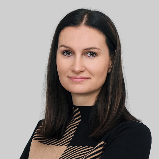 Portret kobiety z ciemnymi włosami, ubranej w czarno-beżową bluzkę w kratę, na jednolitym szarym tle. Uśmiech, profesjonalny wygląd.