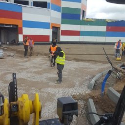 Zubud - Prace przygotowawcze podłoża pod kostkę brukową na terenie inwestycji z kolorową elewacją. Widoczni pracownicy i maszyny budowlane.