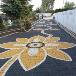 Zubud - Ogród z czarno-żółtą kostką brukową ułożoną we wzór kwiatu i pnącza. W tle biały mur i zieleń. Osoba spryskuje mur. Widok z perspektywy.