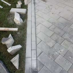 Zubud - Układanie szarej kostki brukowej w jodełkę, widoczne niedocięte elementy na trawie. Krawężnik z kostki oddziela trawnik od ułożonej nawierzchni.