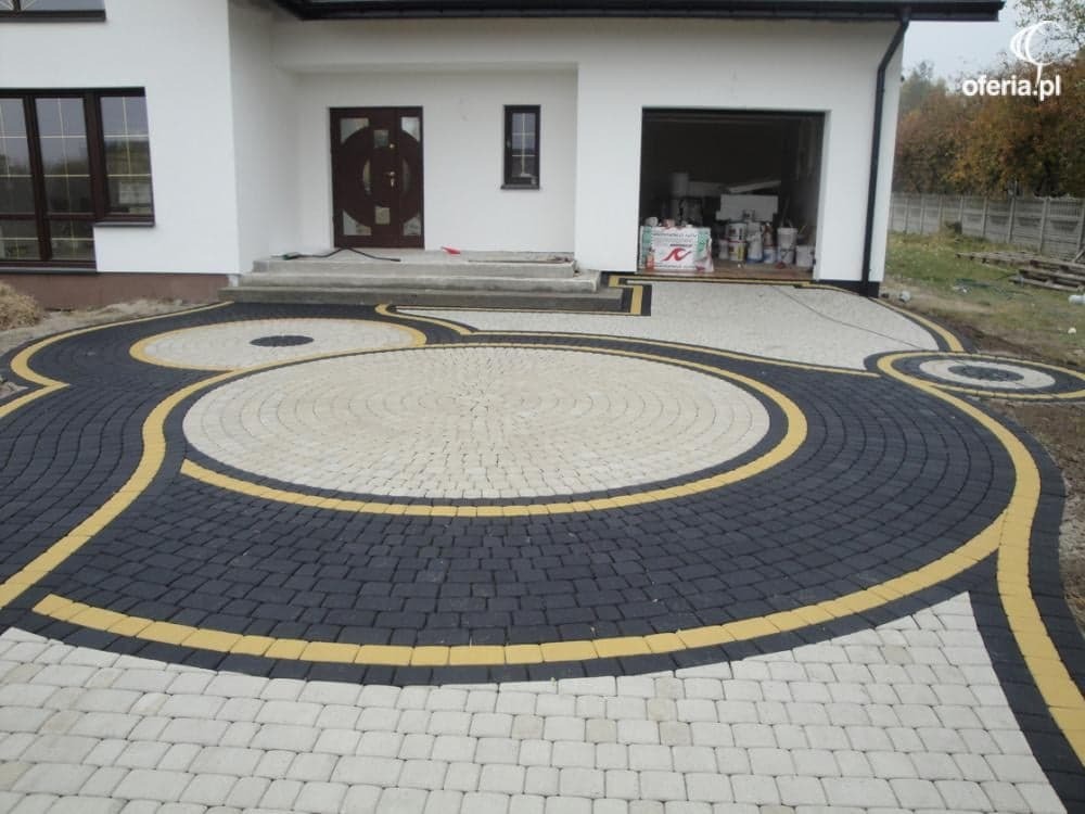 Okrągły wzór z kostki brukowej w odcieniach szarości i beżu, z żółtymi akcentami, prowadzący do białego domu z garażem. Widoczne materiały budowlane w garażu.
