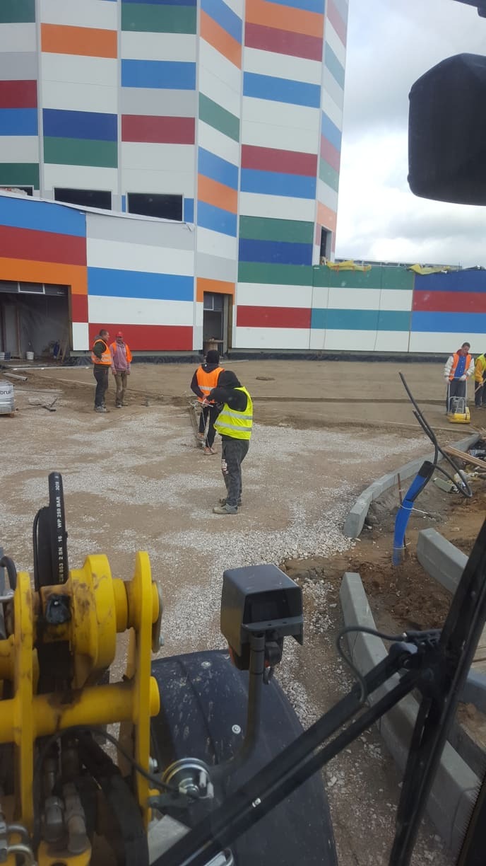 Prace przygotowawcze podłoża pod kostkę brukową na terenie inwestycji z kolorową elewacją. Widoczni pracownicy i maszyny budowlane.