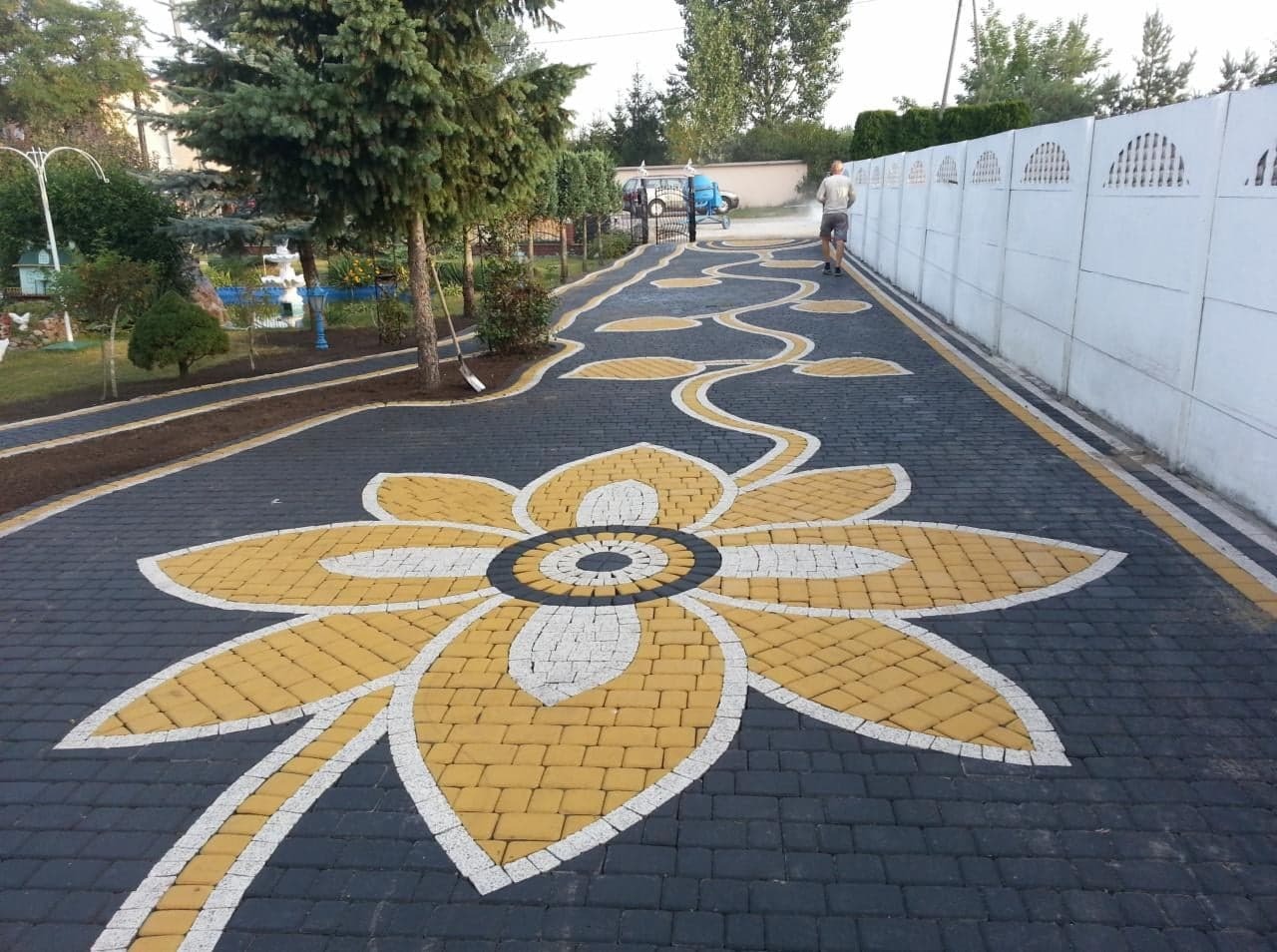 Ogród z czarno-żółtą kostką brukową ułożoną we wzór kwiatu i pnącza. W tle biały mur i zieleń. Osoba spryskuje mur. Widok z perspektywy.