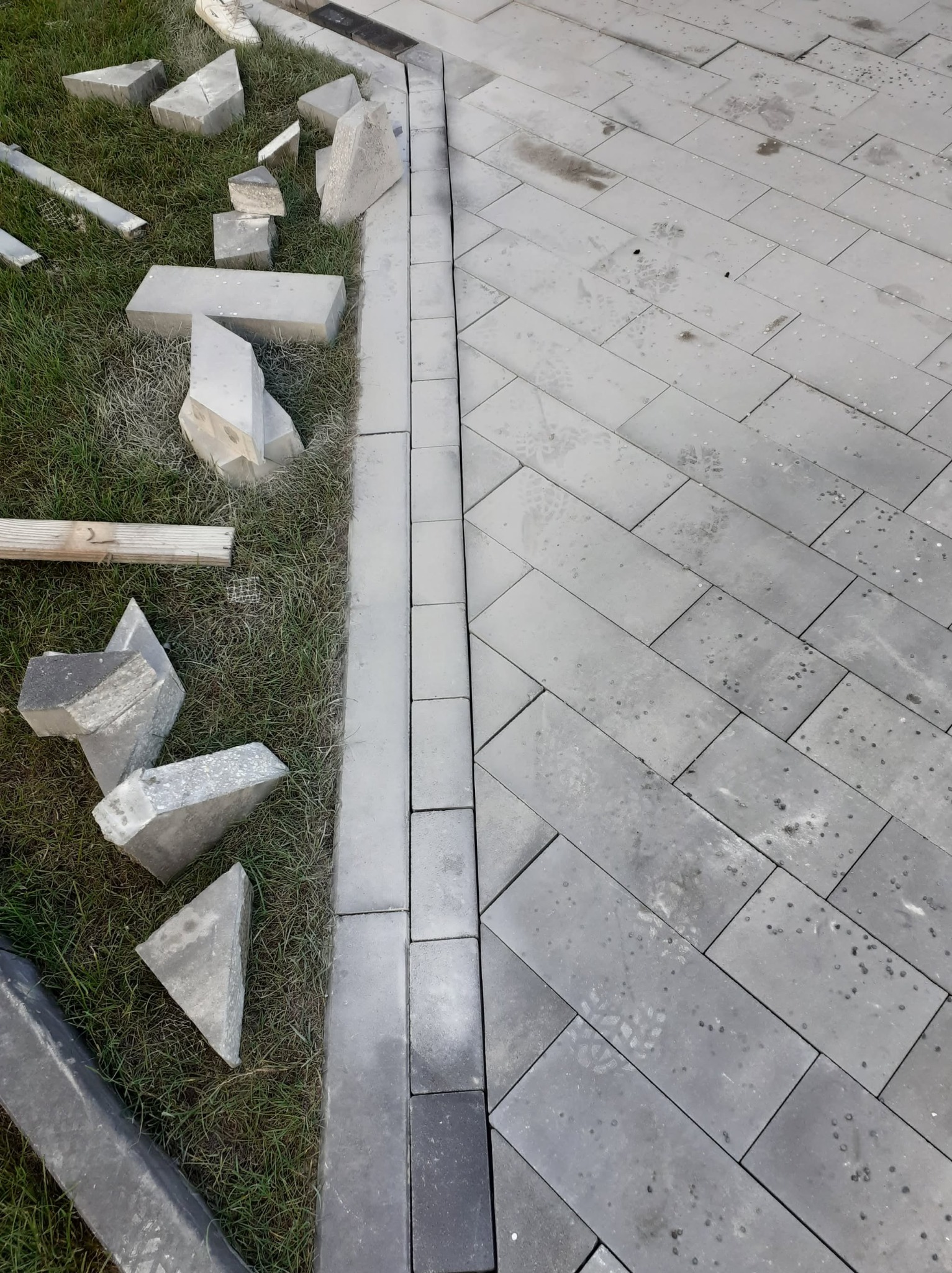 Układanie szarej kostki brukowej w jodełkę, widoczne niedocięte elementy na trawie. Krawężnik z kostki oddziela trawnik od ułożonej nawierzchni.