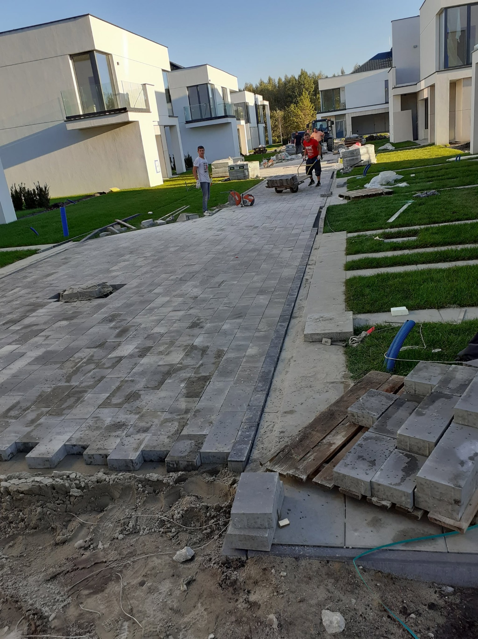 Układanie szarej kostki brukowej na posesji z nowoczesnymi domami. Widoczny fragment ułożonej nawierzchni, paleta z kostką i pracownicy w tle.